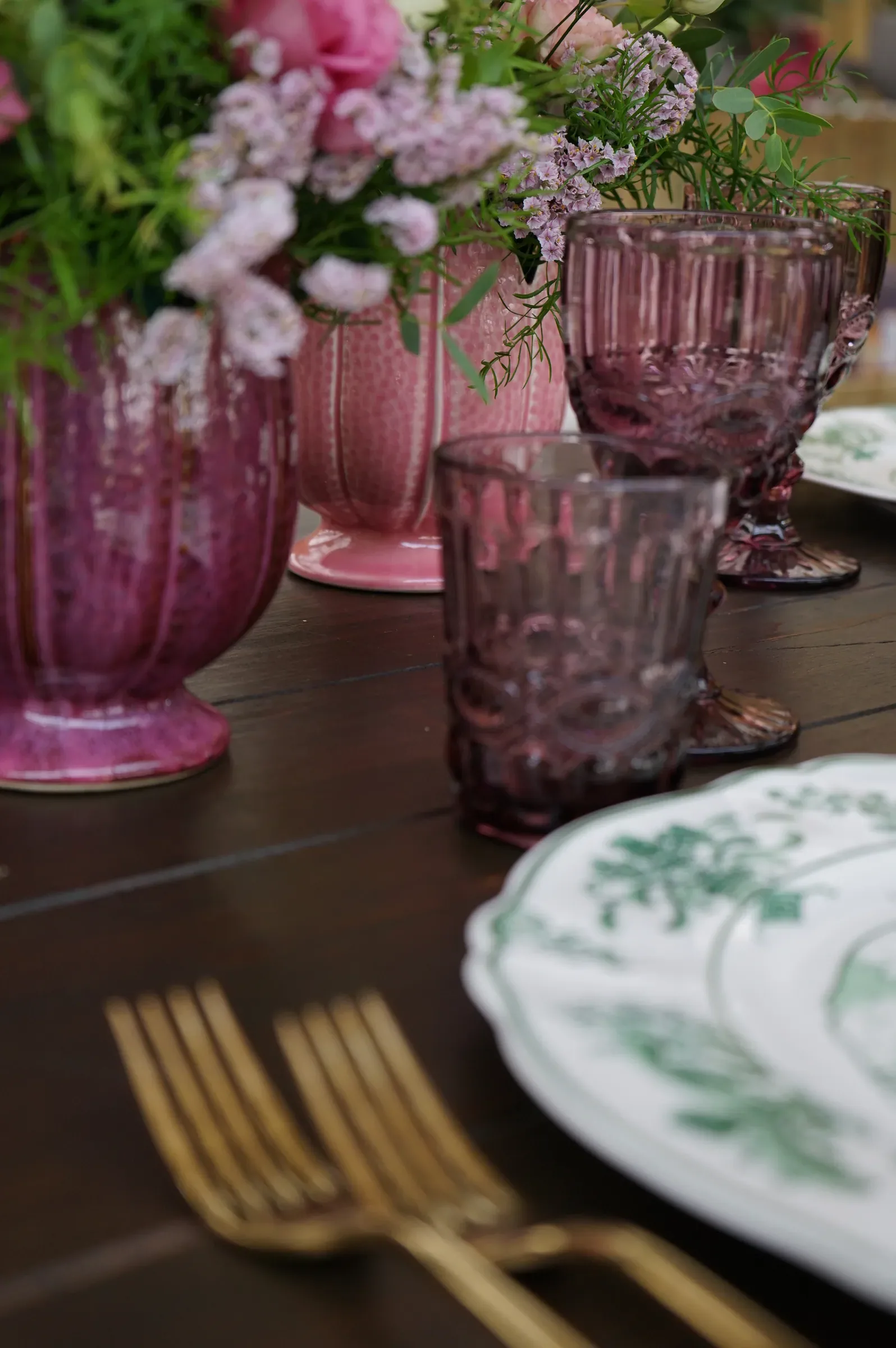 Vasi di ceramica rosa e calici di vetro viola accanto al piatto a decoro verde, con le forchette dorate