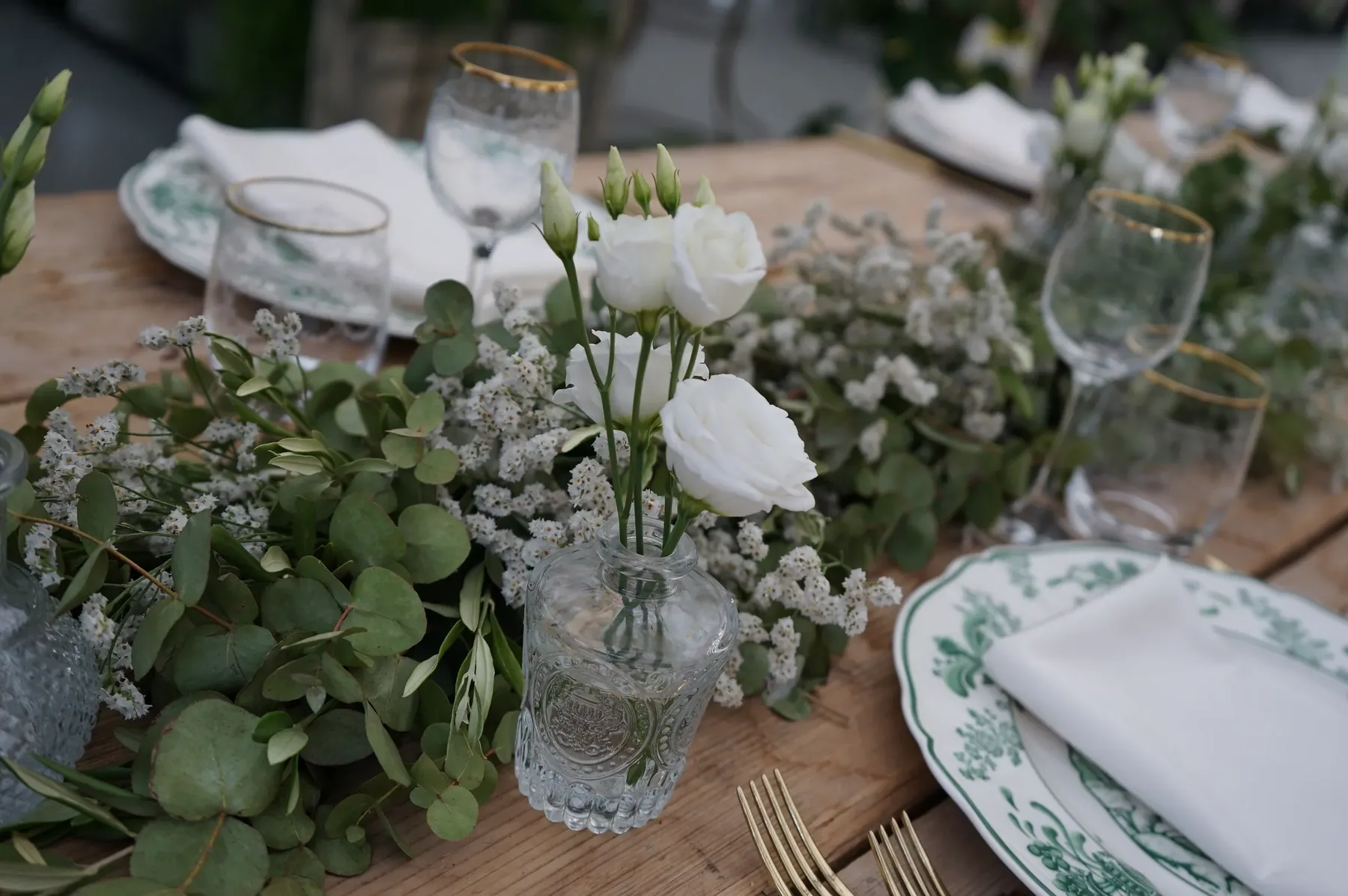 Rose bianche, eucalipto e fiori bianchi in vasetti di vetro lungo il tavolo, accanto al piatto a decoro verde