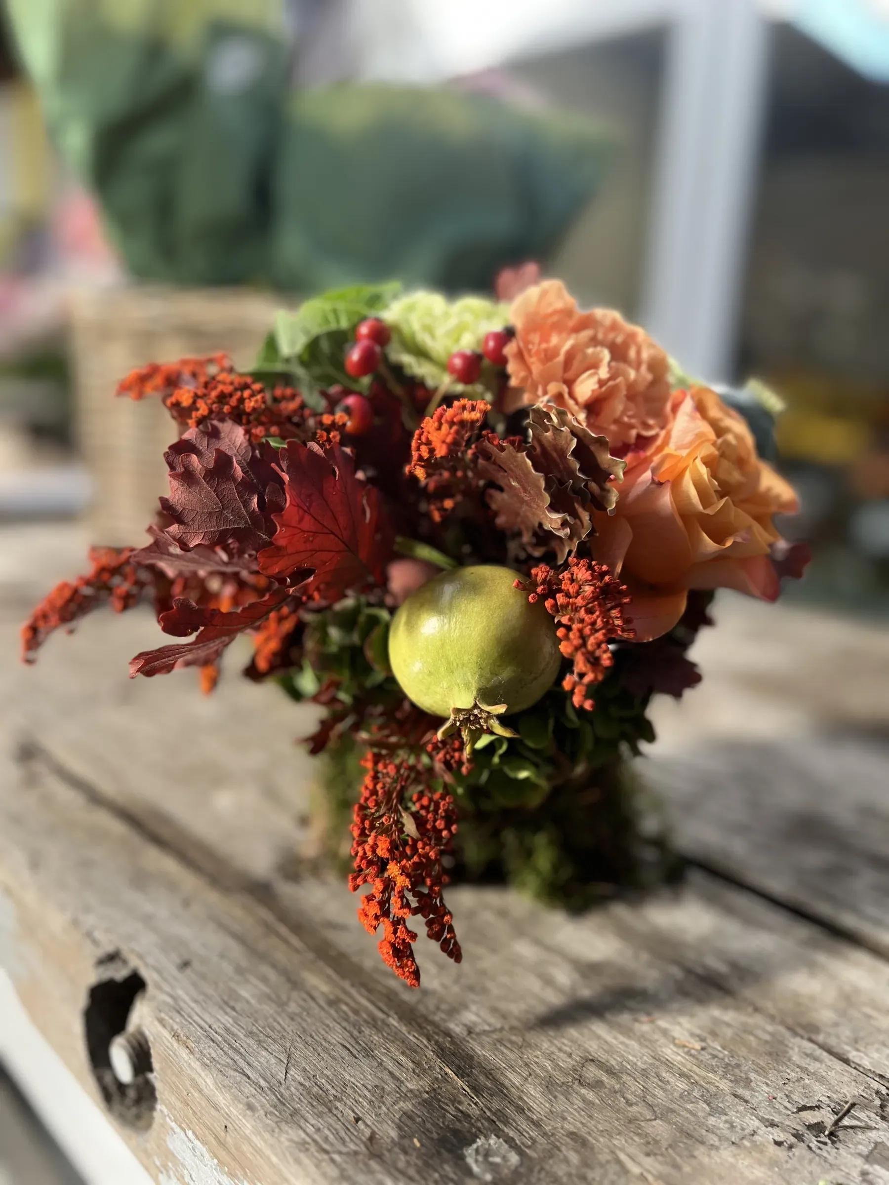 Composizione bassa di rose arancio, foglie rosse, bacche e un frutto verde in un vaso rivestito di muschio, su un tavolo di legno