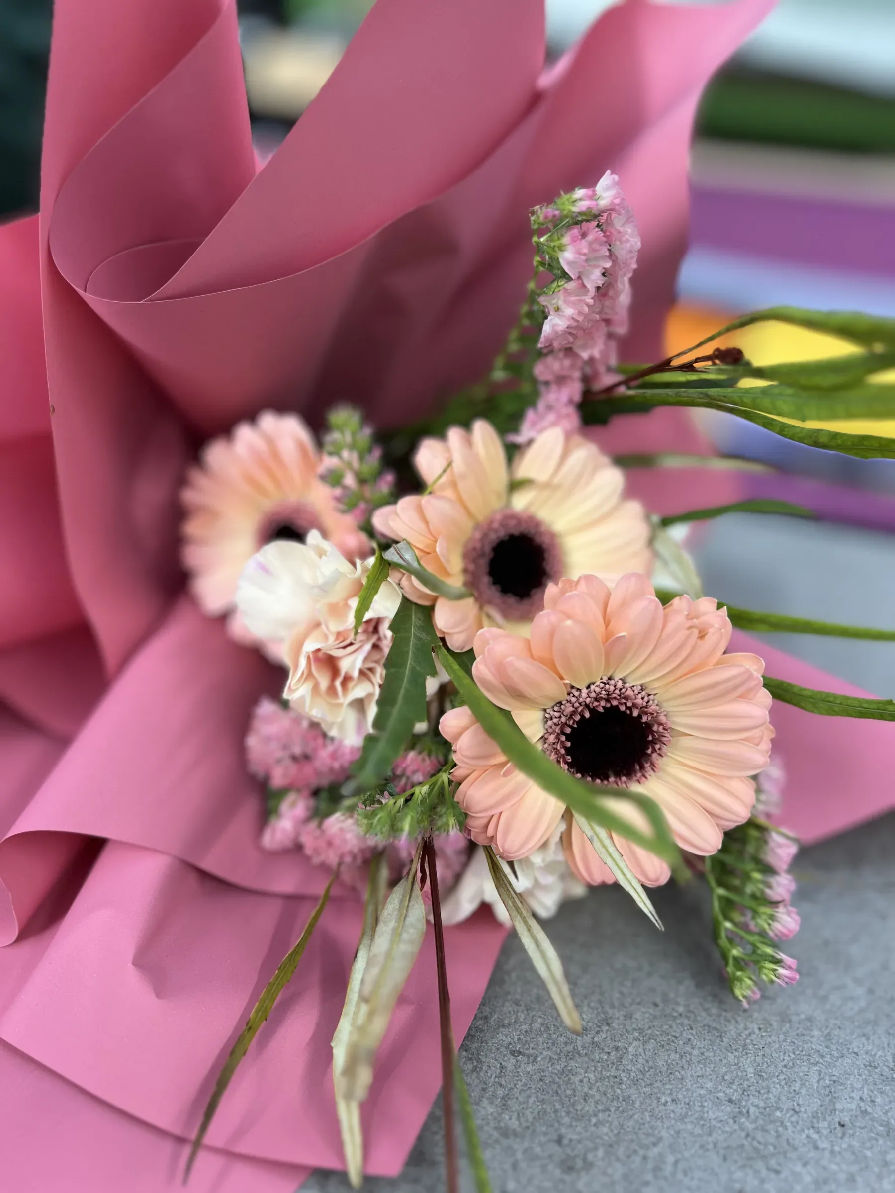 Mazzo di gerbere color pesca, garofani chiari e limonium rosa, avvolto nella carta rosa e appoggiato sul banco