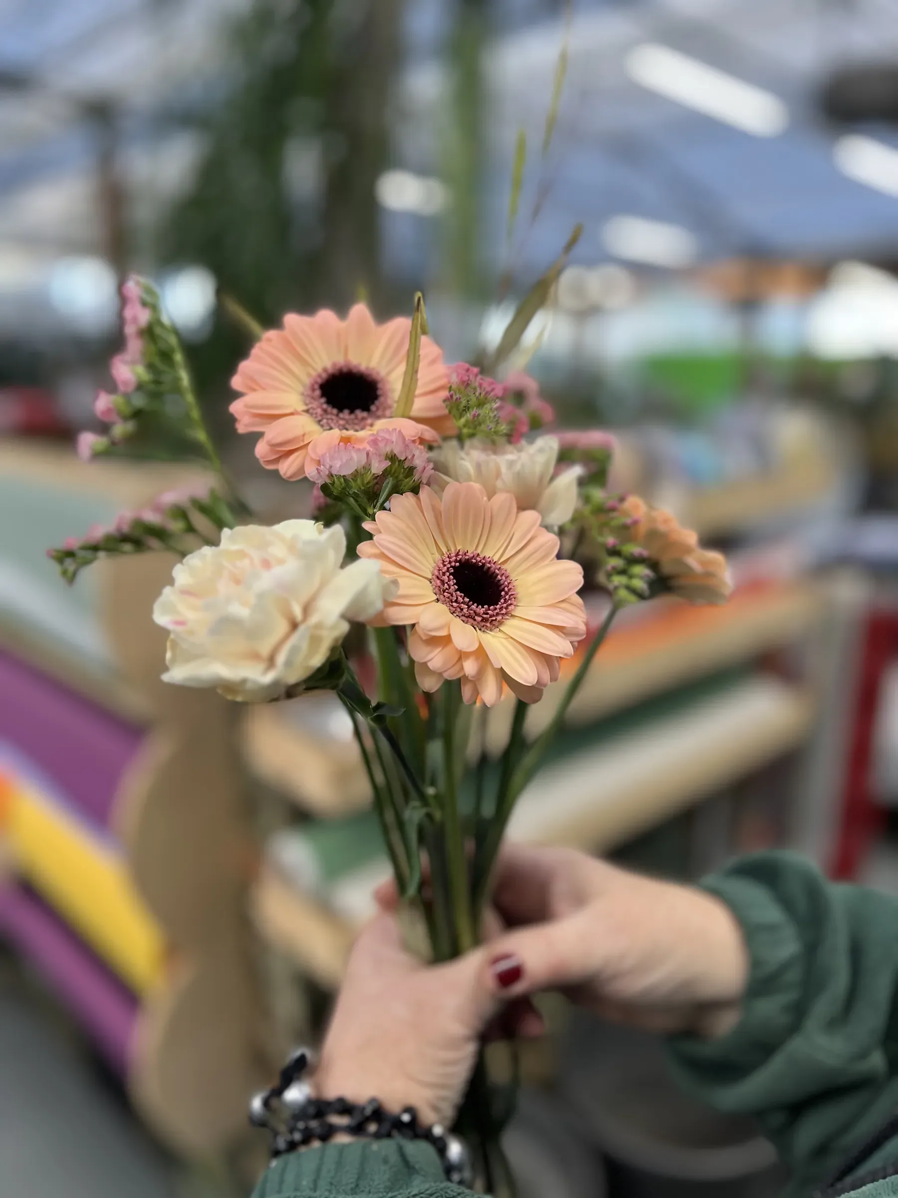 Mazzolino di gerbere color pesca, garofani chiari e limonium rosa tenuto in mano davanti agli scaffali della carta