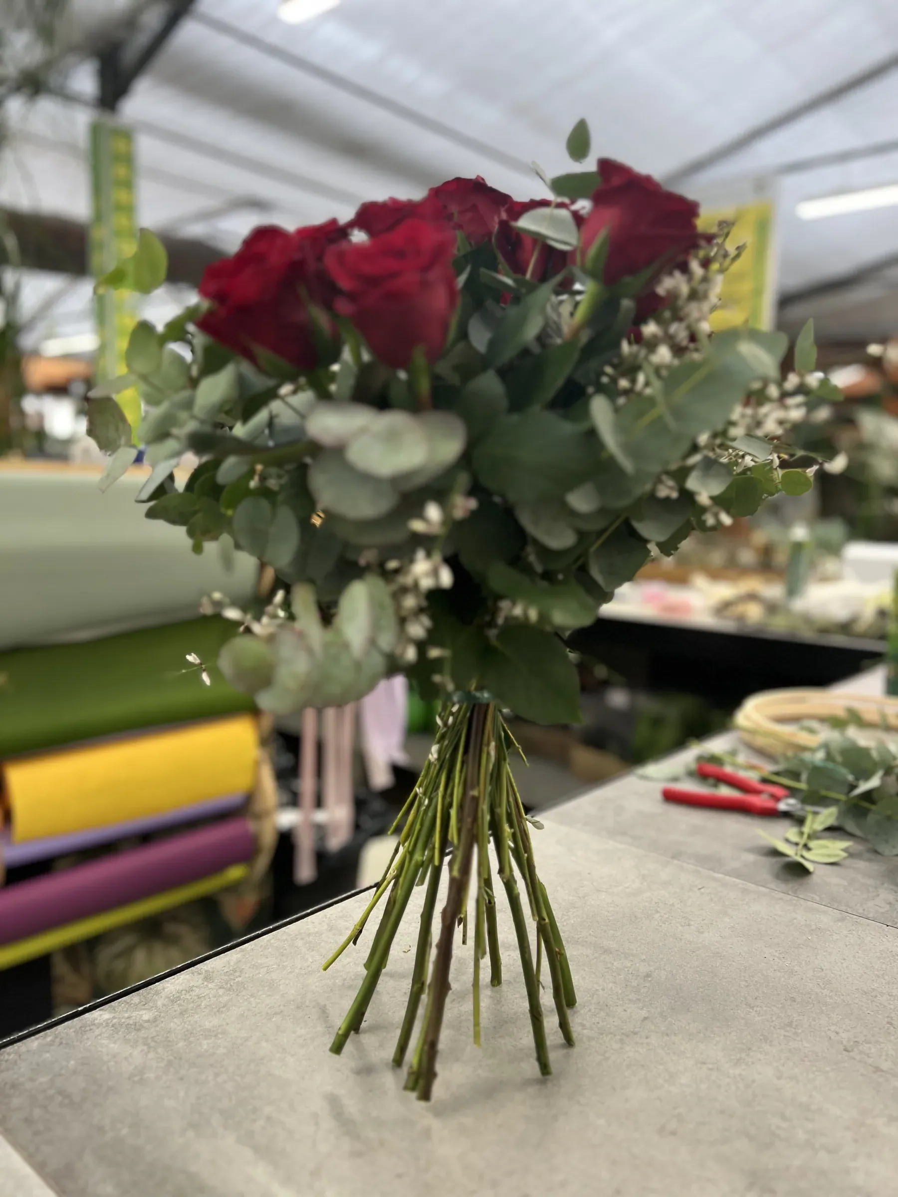 Mazzo di rose rosse con eucalipto e fiorellini bianchi, dritto sul banco accanto ai rotoli di carta e alle forbici