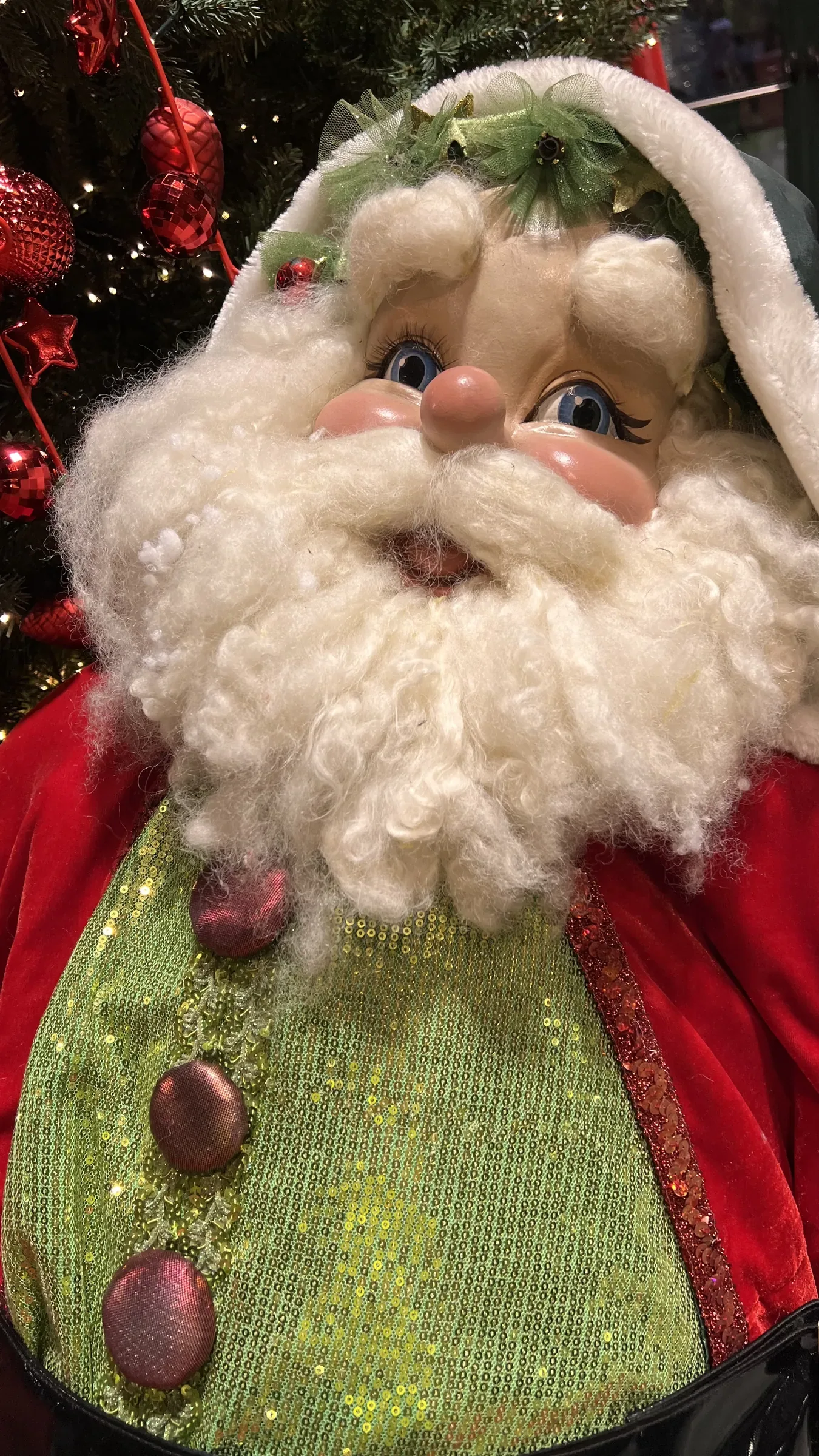 Pupazzo di Babbo Natale da vicino, con la barba di lana e il gilet verde di paillettes, davanti a un albero addobbato di rosso