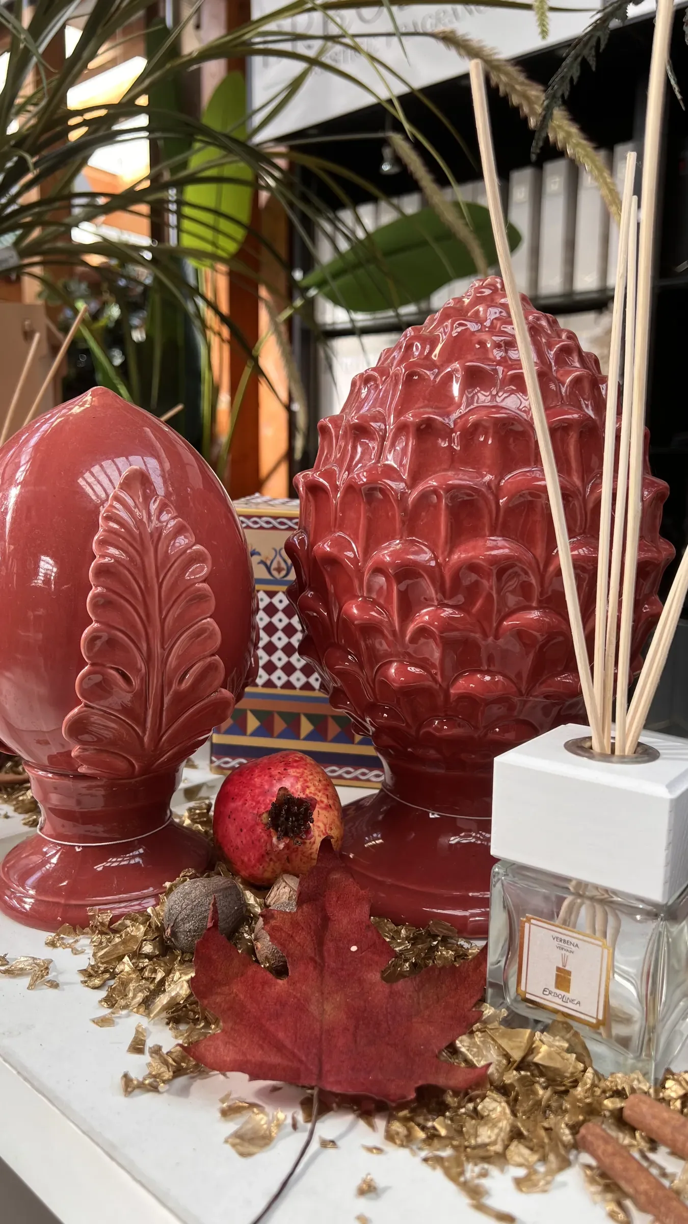 Una pigna e un pumo in ceramica rossa accanto a un diffusore a bastoncini, con una melagrana e una foglia d'acero rossa