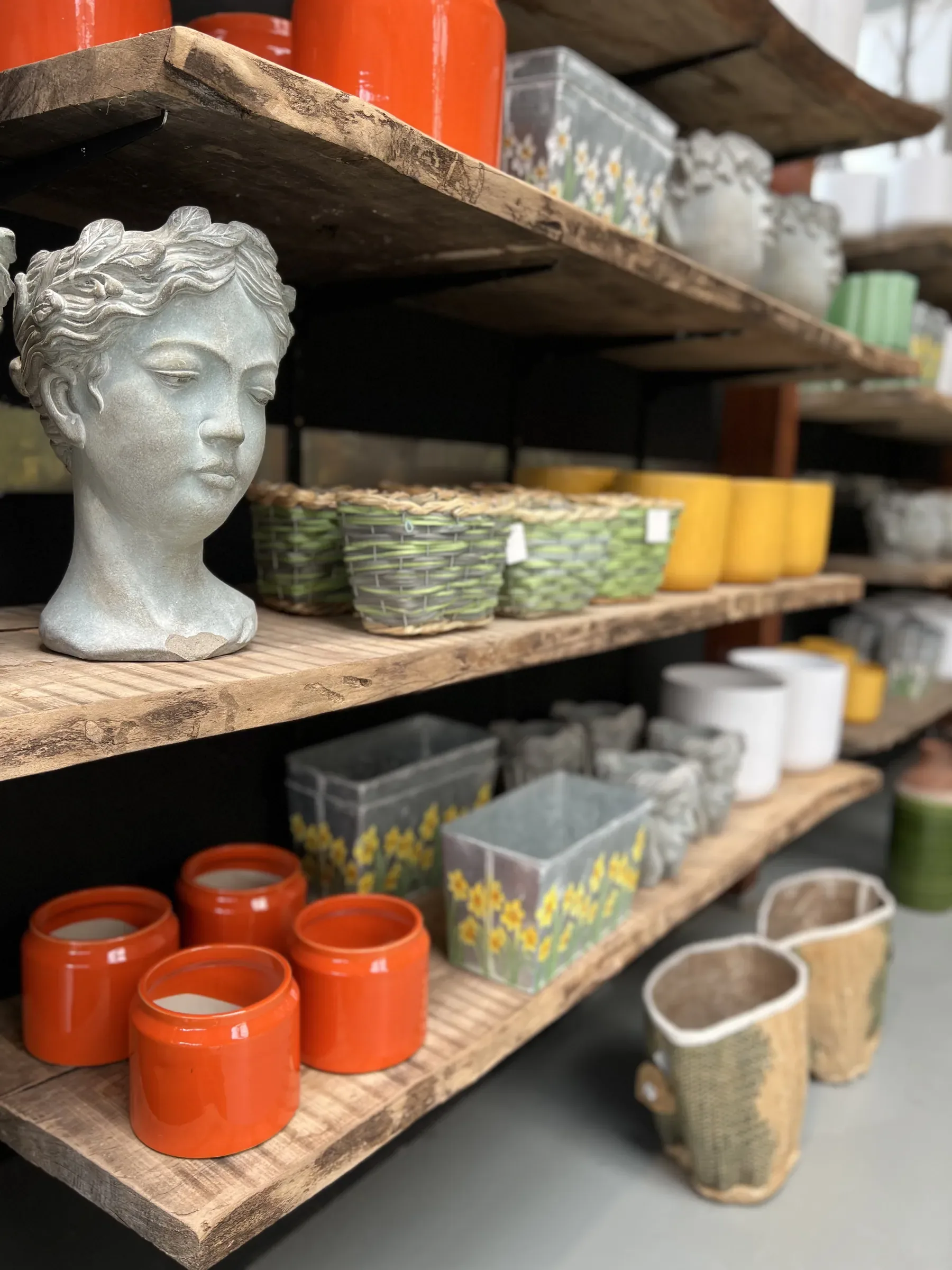 Scaffali in legno grezzo con vasi smaltati arancioni, cestini intrecciati, un vaso a forma di testa e contenitori di zinco dipinti a narcisi