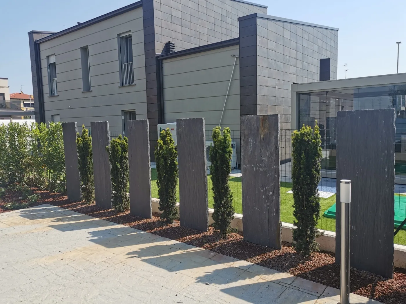 Fila di lastre verticali in ardesia alternate a cipressi colonnari, lungo un camminamento in pietra grigia