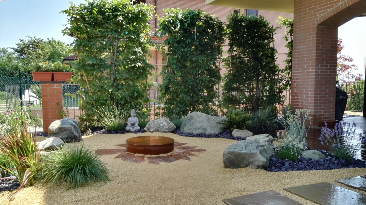 Cortile d'ingresso con grigliati coperti di rampicanti, vasca circolare in acciaio corten posata sulla ghiaia, massi e graminacee ai lati