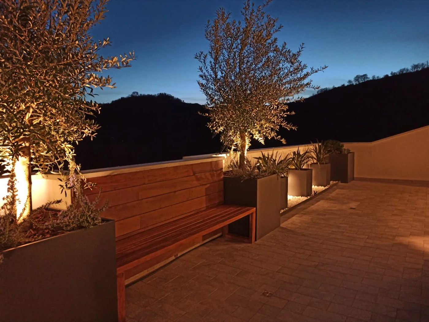 Terrazzo di sera con un albero illuminato dal basso in una fioriera, la panca in legno e la collina scura sullo sfondo