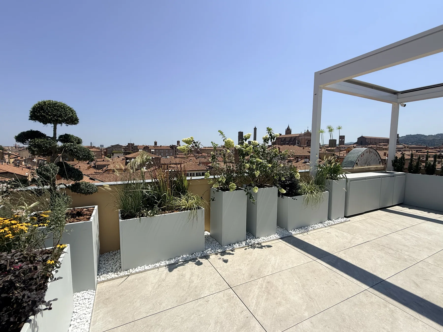 Terrazzo sui tetti della città con fioriere alte chiare, alberelli potati a nuvola e la struttura bianca di una pergola