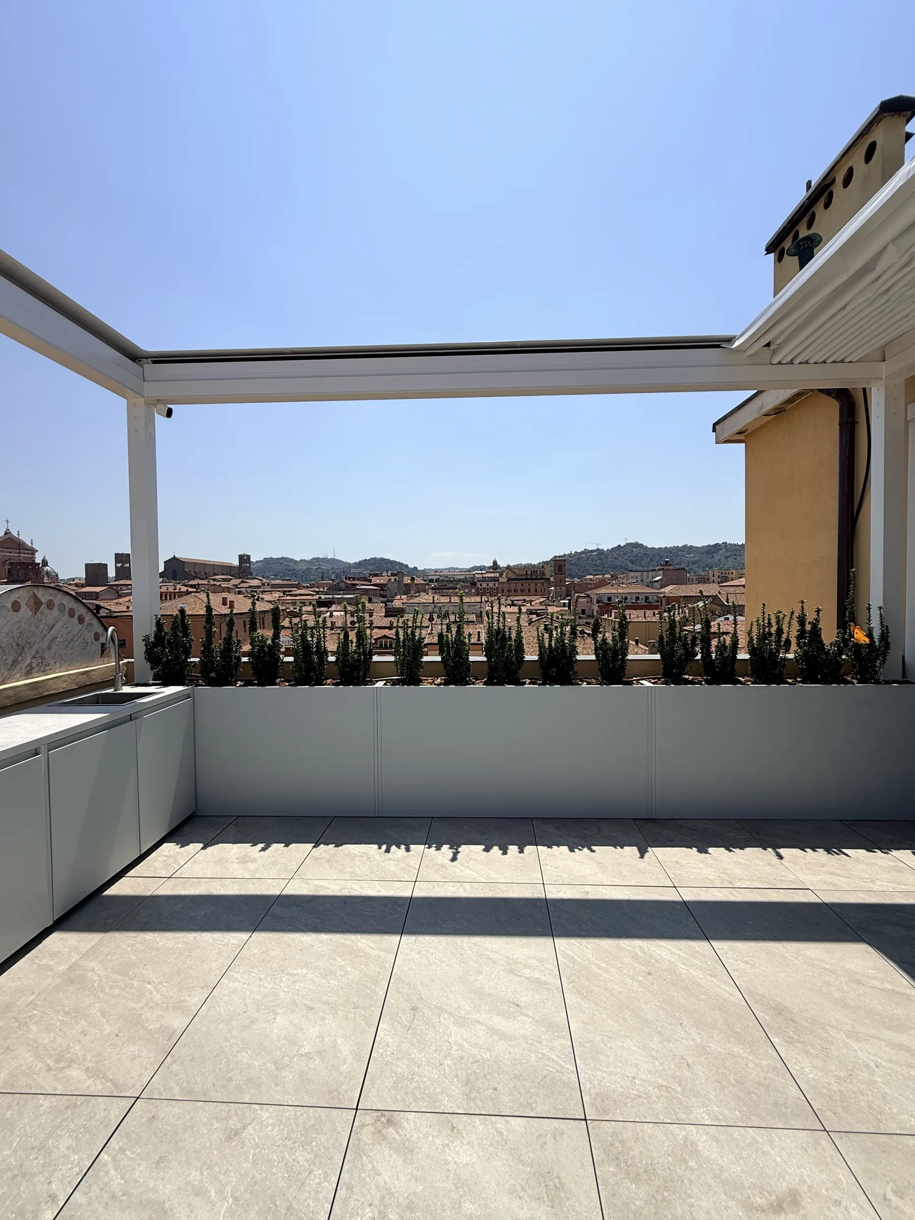 Lo stesso terrazzo dopo il lavoro, sotto la pergola: pavimento in gres chiaro, una cucina da esterno e fioriere alte con piccoli cipressi lungo il parapetto