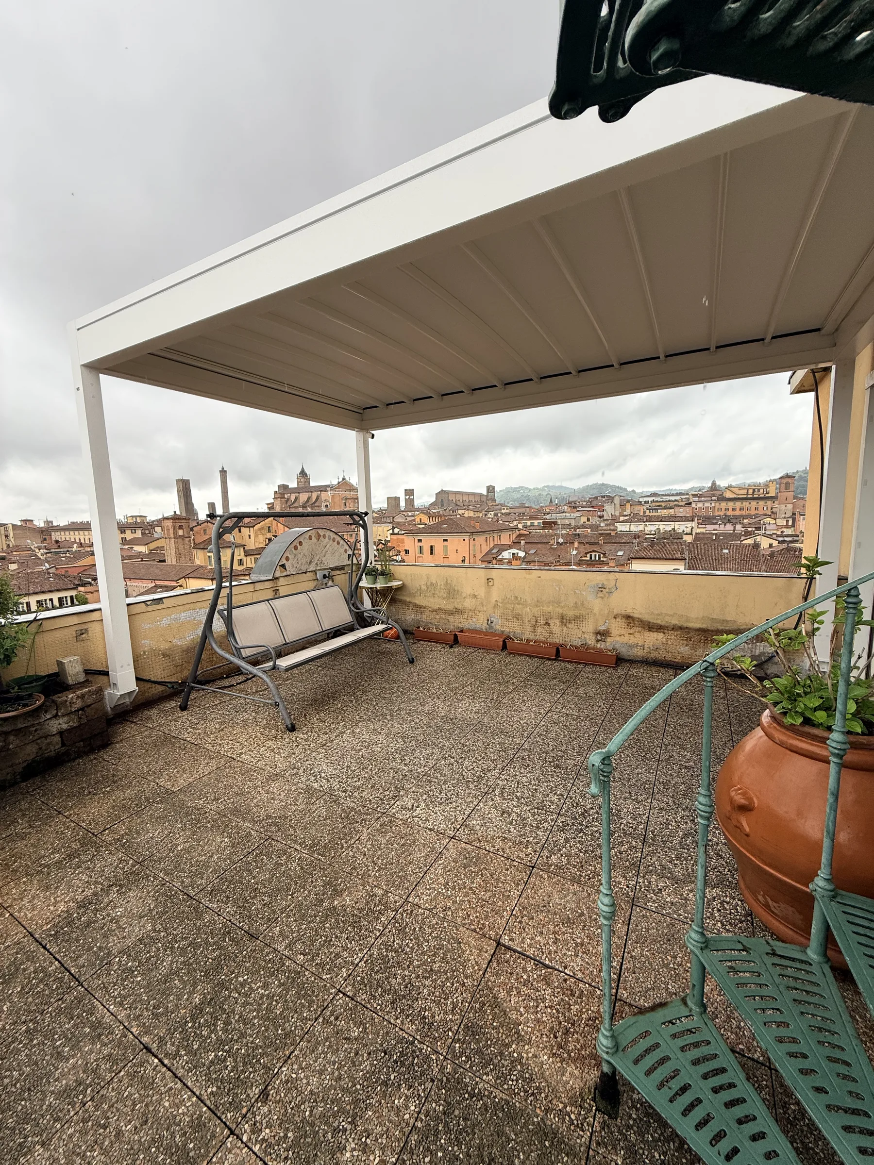 Il terrazzo prima del lavoro, sotto la pergola: pavimento in graniglia consumato, un dondolo e un vaso di terracotta, i tetti di Bologna sullo sfondo
