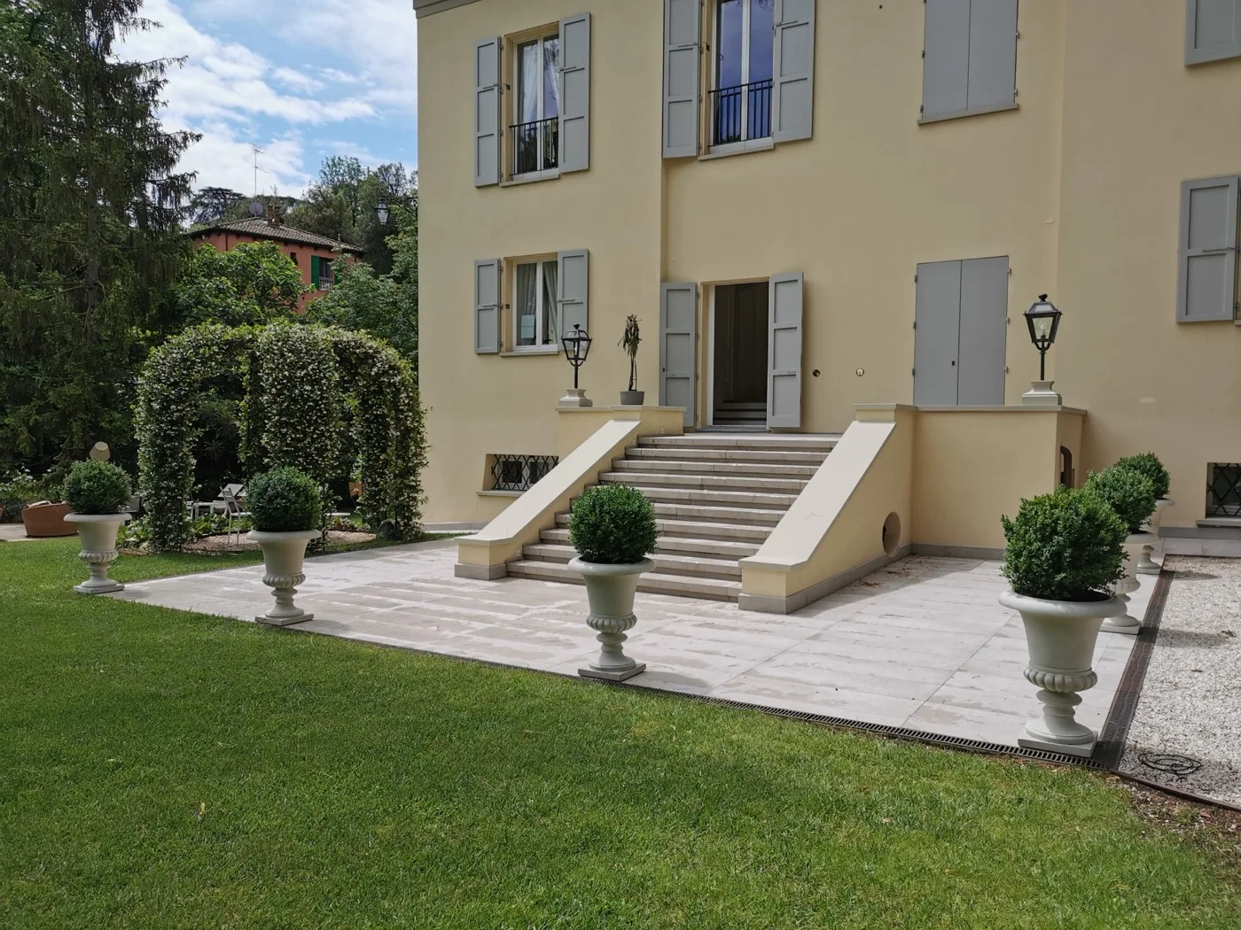 Fronte di una villa con scalinata in pietra, arco di gelsomino potato, vasi in cemento con sfere di bosso e prato rasato attorno