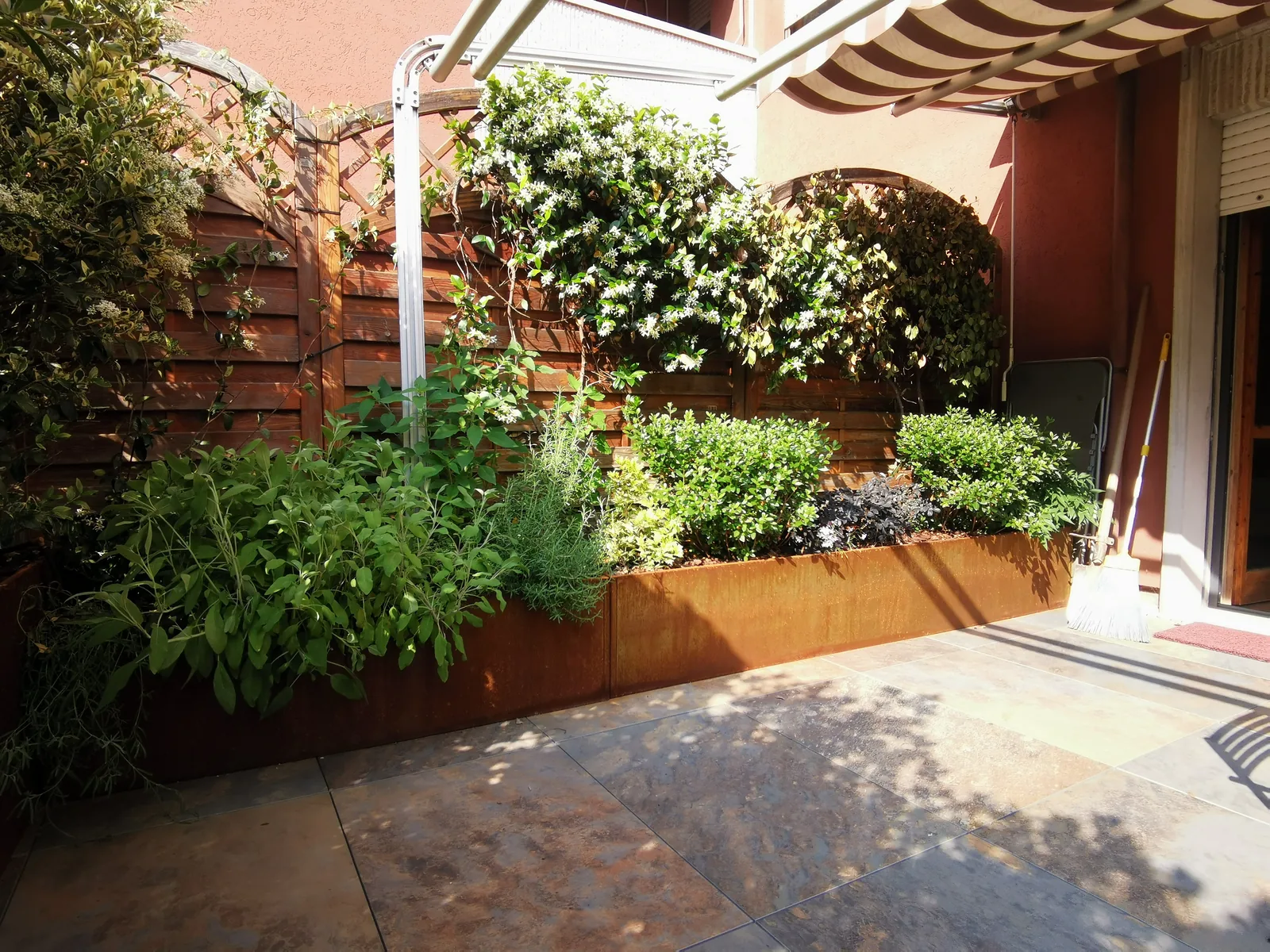 Fioriera in corten ad angolo con arbusti e aromatiche, sotto i rampicanti sul grigliato in legno