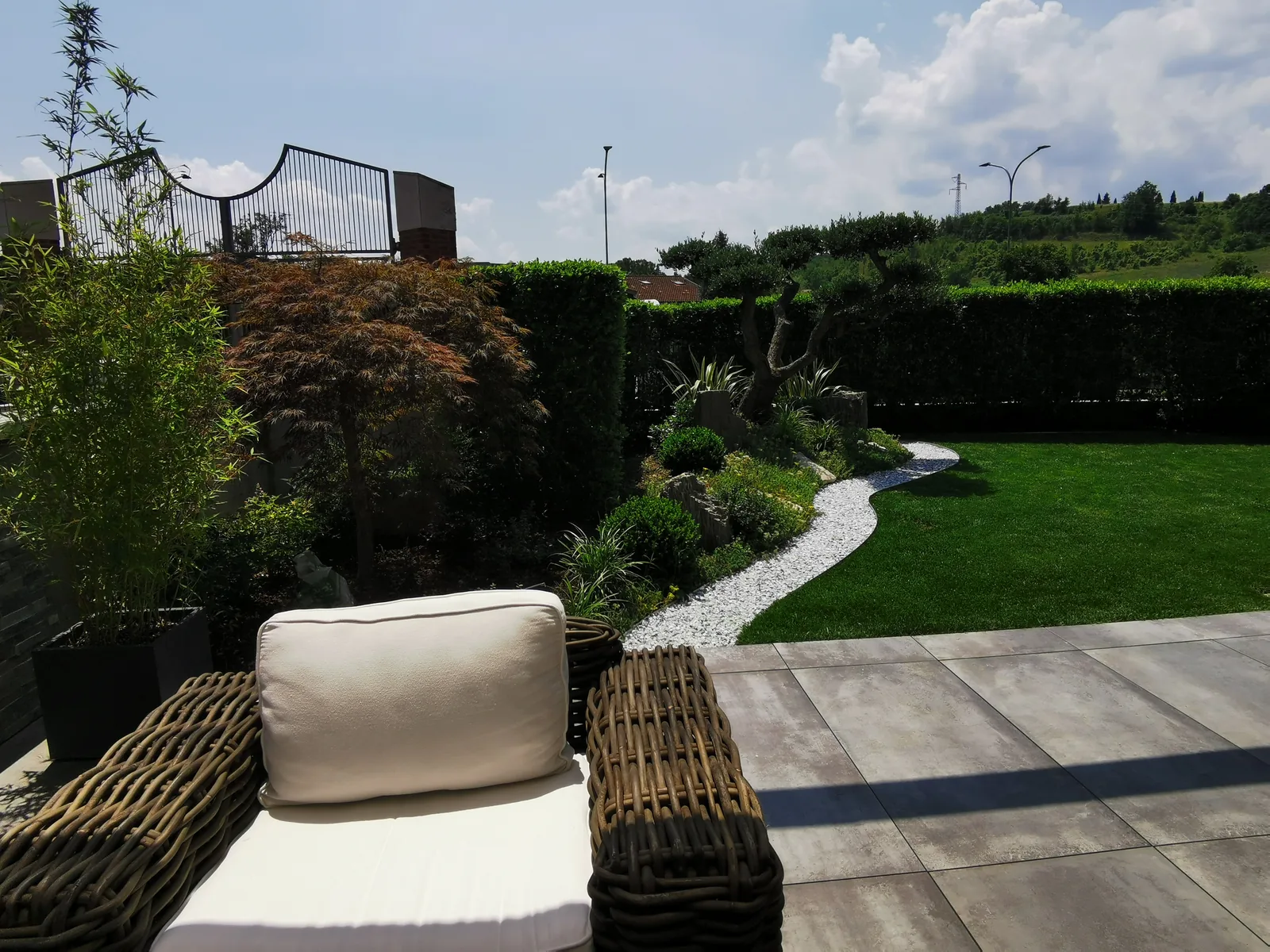 Poltrona in rattan con cuscino chiaro sulla pavimentazione in lastre, con il prato e il vialetto bianco sul fondo
