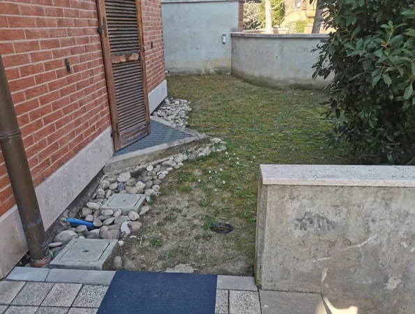 Prima: angolo del giardino con prato rado, sassi lungo il muro in mattoni e un muretto in cemento