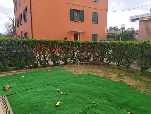 Prima: telo verde sul terreno lungo la siepe, con la casa arancione sul fondo