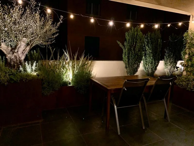 Il terrazzo di sera, con le lucine accese sopra il tavolo, l'ulivo illuminato e le fioriere