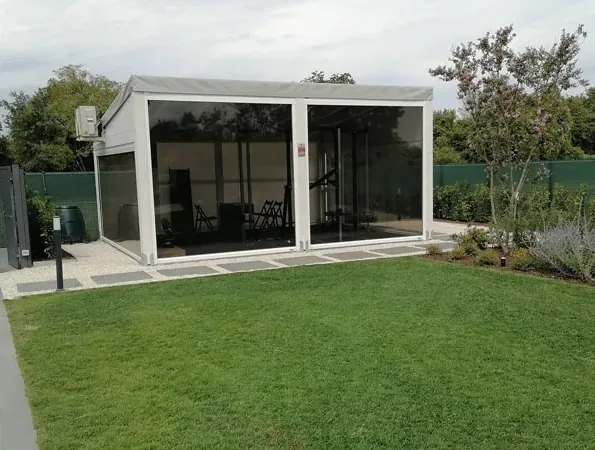 Dopo: pergola bianca chiusa da vetrate sul prato, con un alberello e arbusti accanto