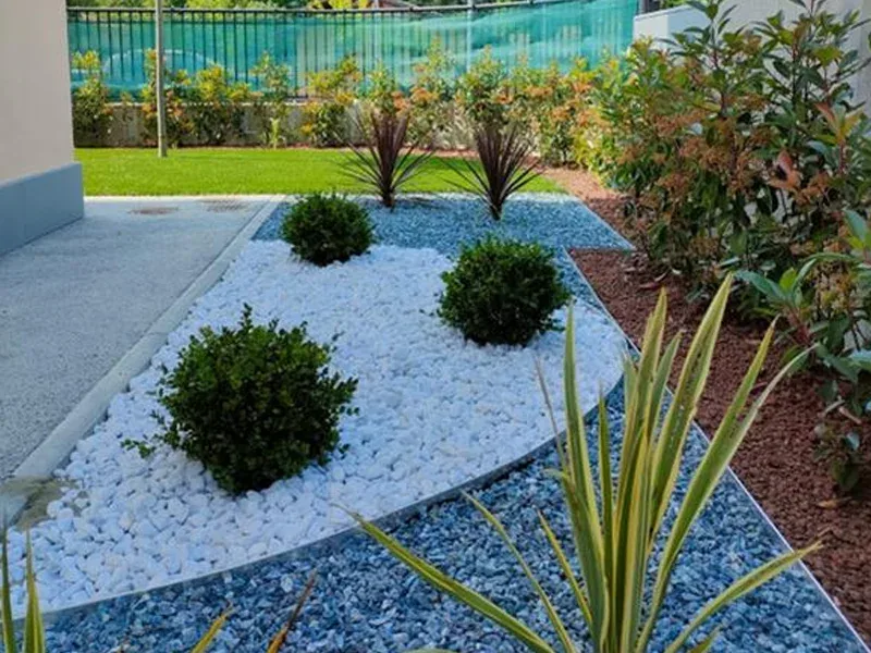 Aiuola d'ingresso con ghiaia bianca e azzurra, sfere di bosso e phormium, con il prato sintetico e la siepe di photinia sul fondo