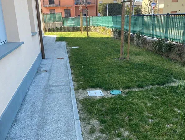 Prima: striscia di prato naturale rado fra il vialetto in cemento della casa e la recinzione verde