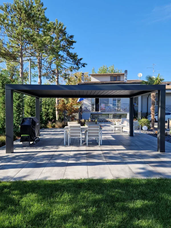 Pergola bioclimatica con tavolo e sedie bianche su lastre grigie, con il prato in primo piano e i pini dietro