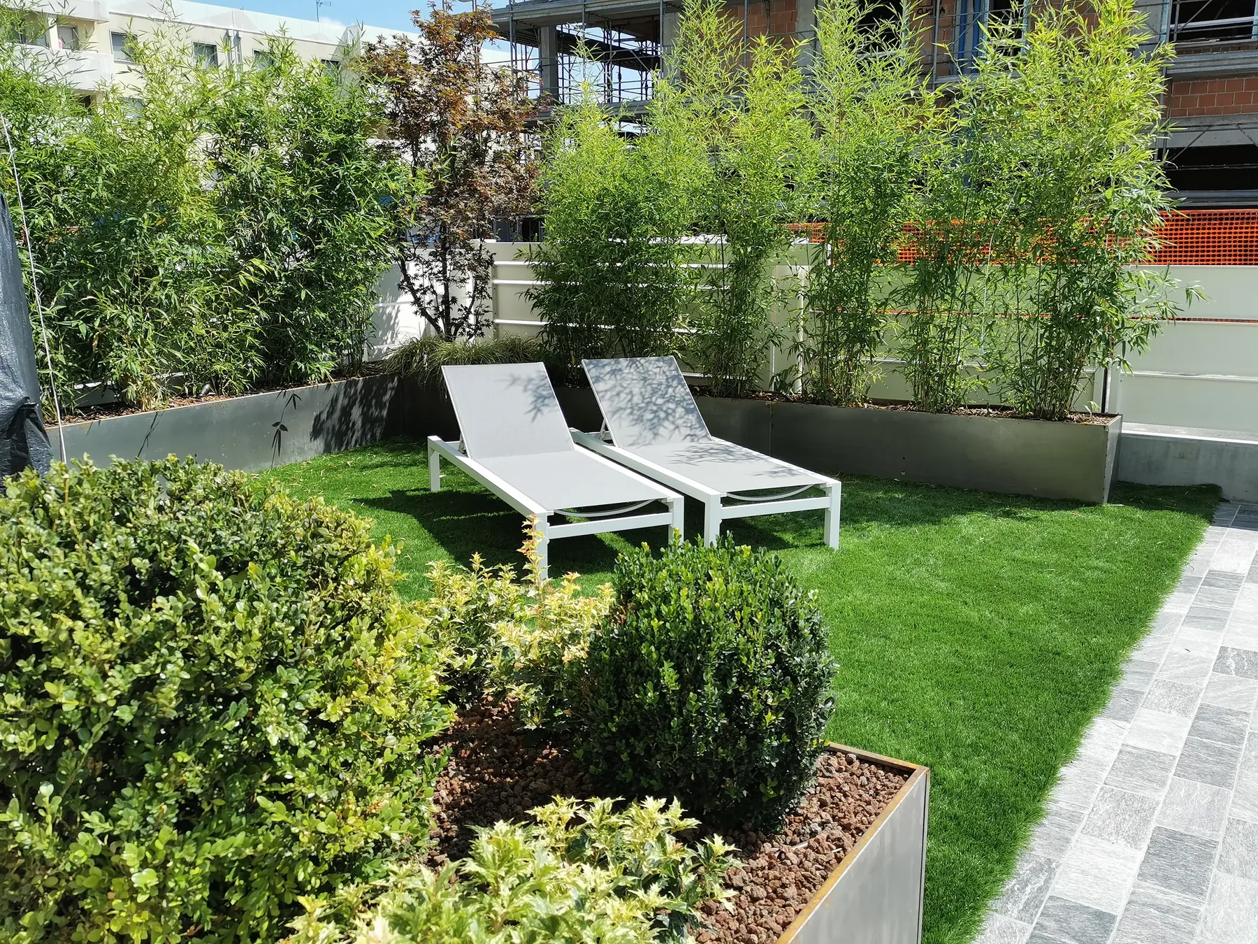 Giardino pensile con prato sintetico e lettini bianchi, chiuso da fioriere di bambù, con sfere di bosso in primo piano
