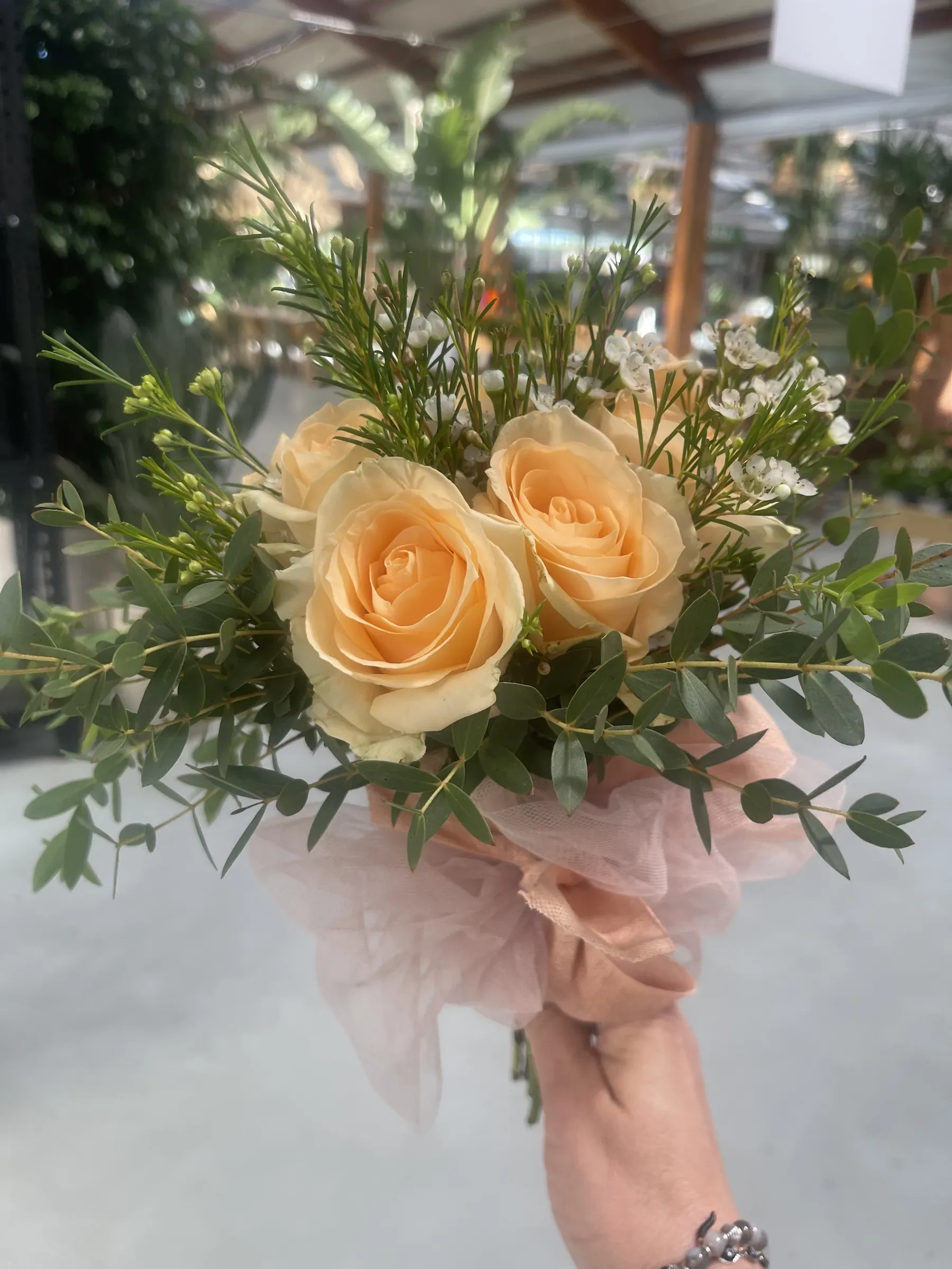 Mazzo di rose color pesca con verde ornamentale e piccoli fiori bianchi, incartato in tulle rosa e tenuto in mano