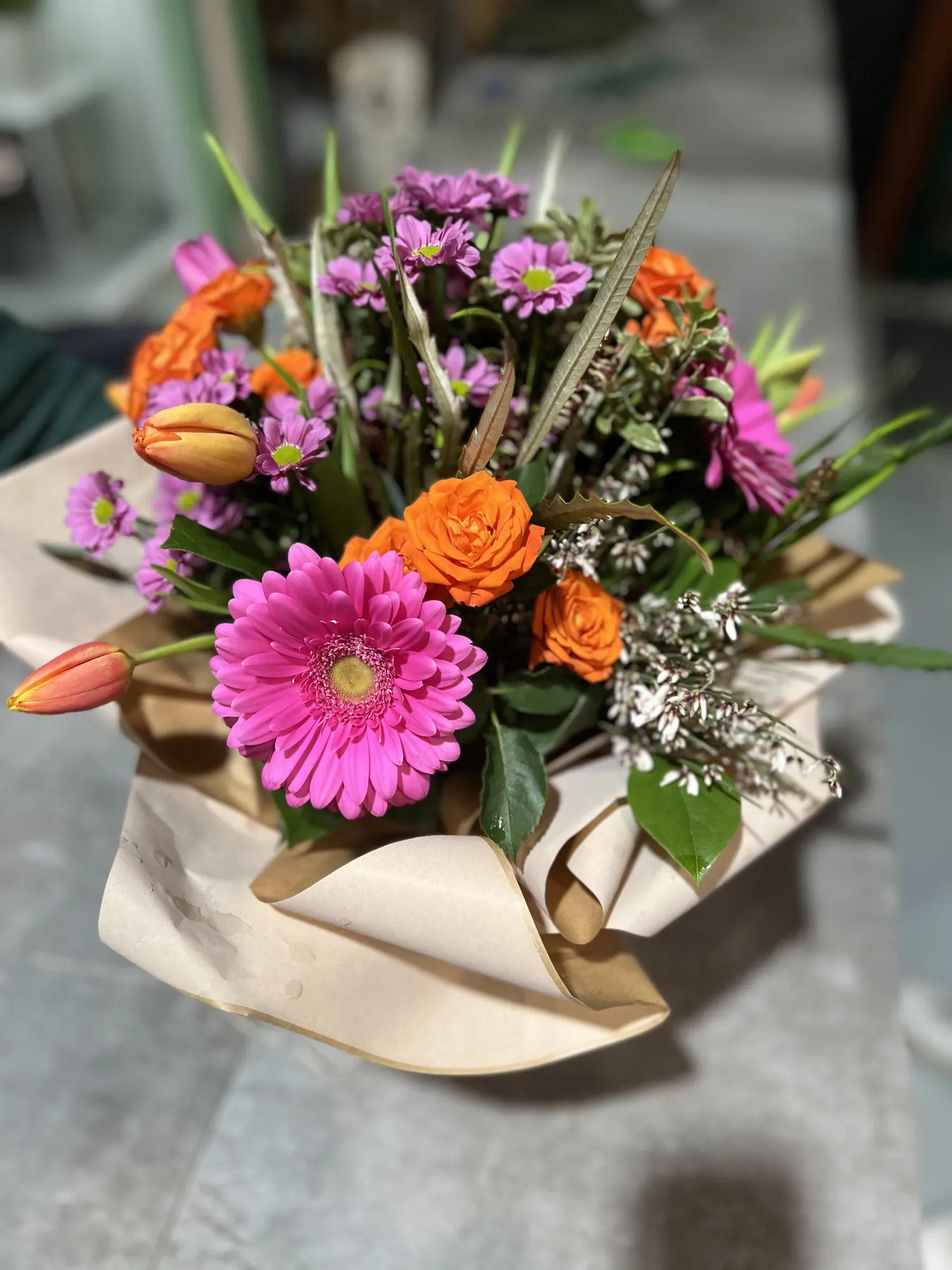 Composizione di gerbere fucsia, rose arancio, tulipani e margherite viola in una confezione di carta color crema