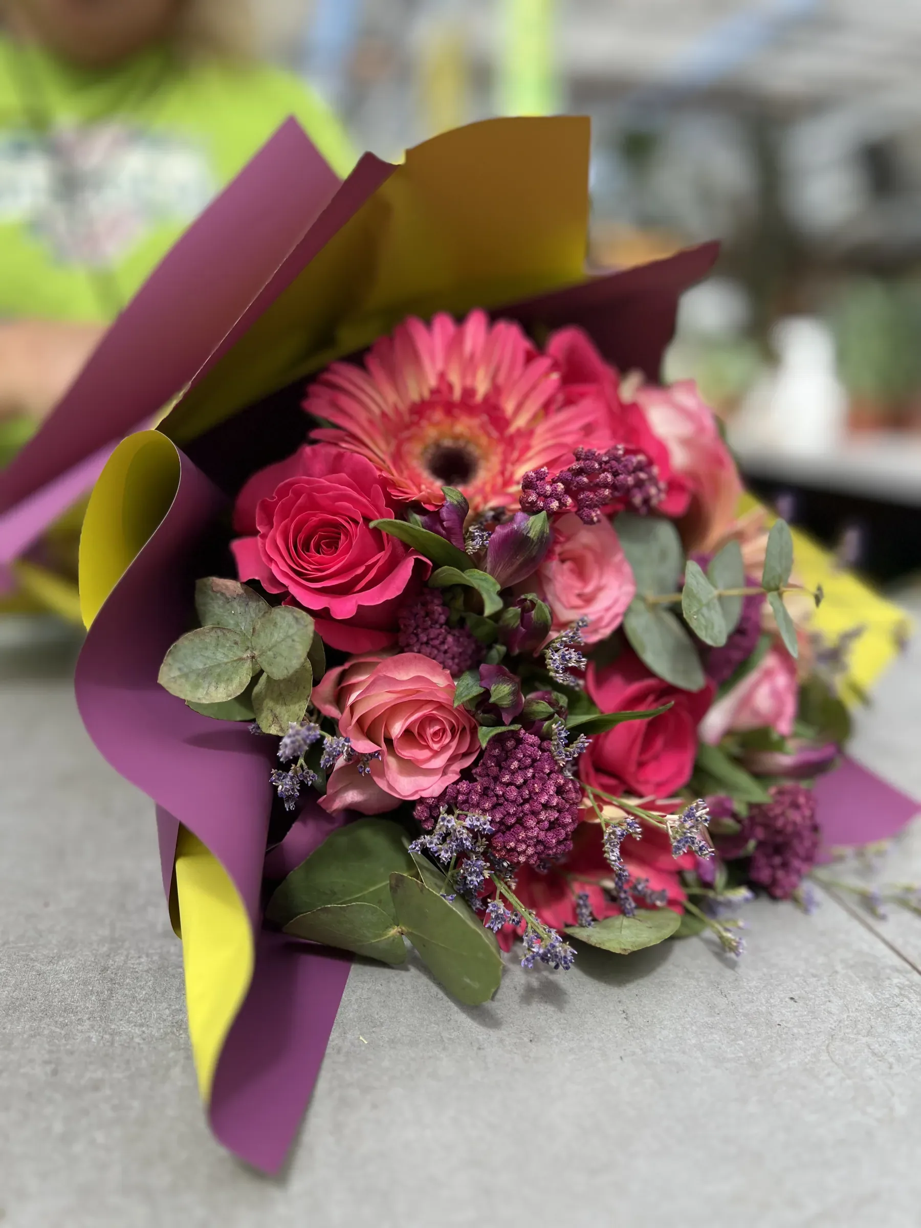 Mazzo di gerbere e rose rosa e fucsia con verde ornamentale, incartato in carta viola e gialla