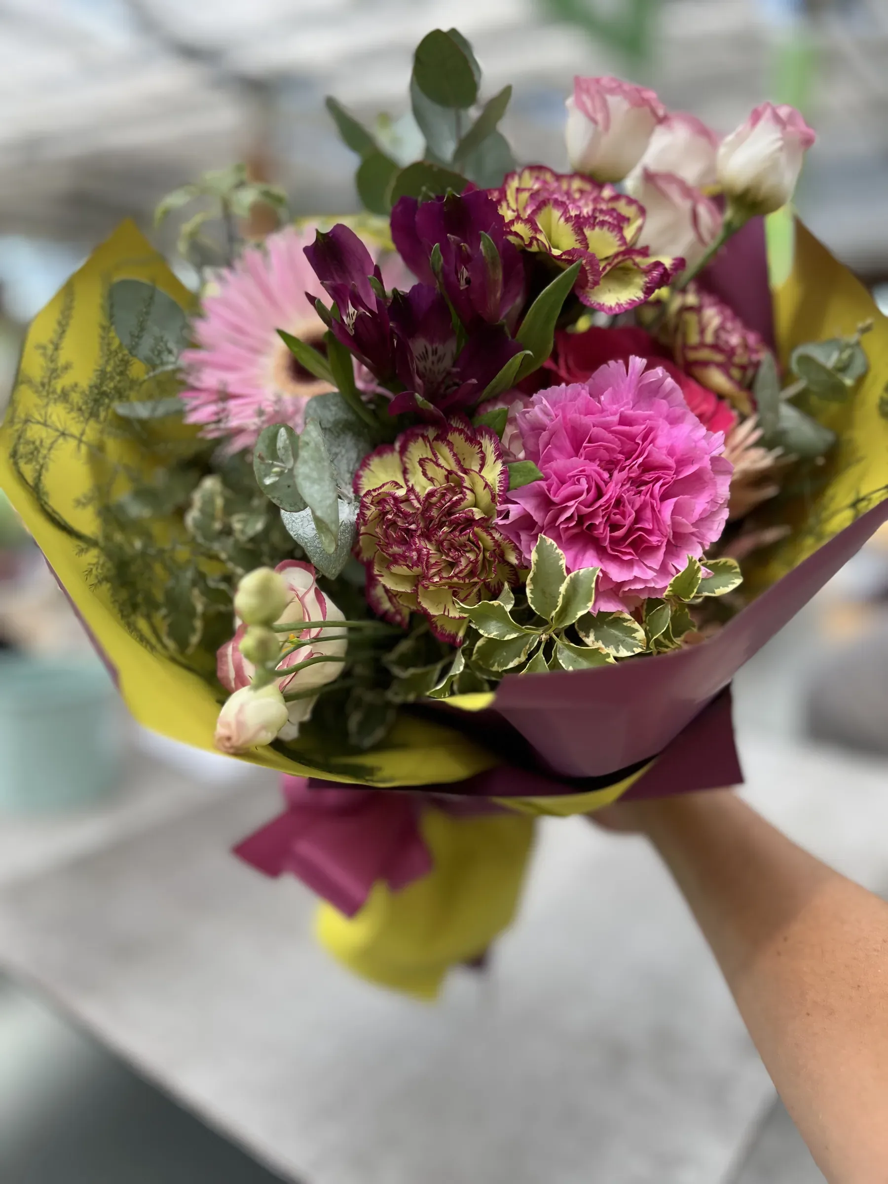 Mazzo di garofani sfumati, gerbera rosa e rose chiare in carta gialla e viola, tenuto in mano