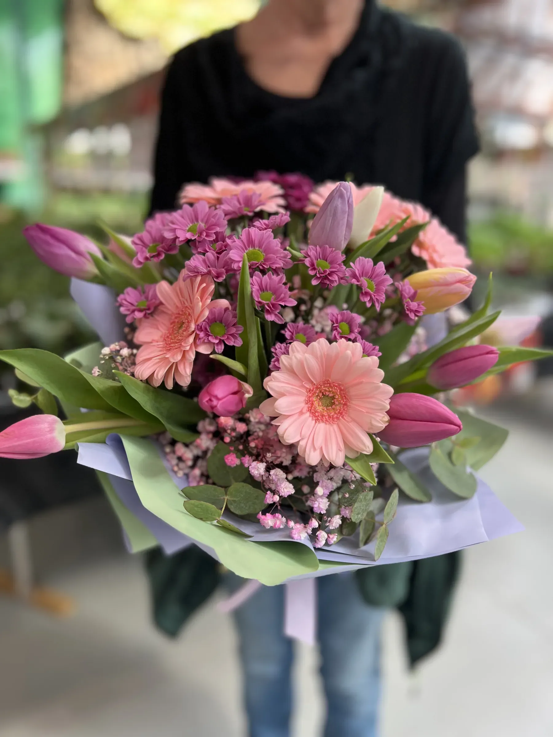 Mazzo di gerbere rosa, tulipani e margherite fucsia in carta lilla, tenuto davanti a sé