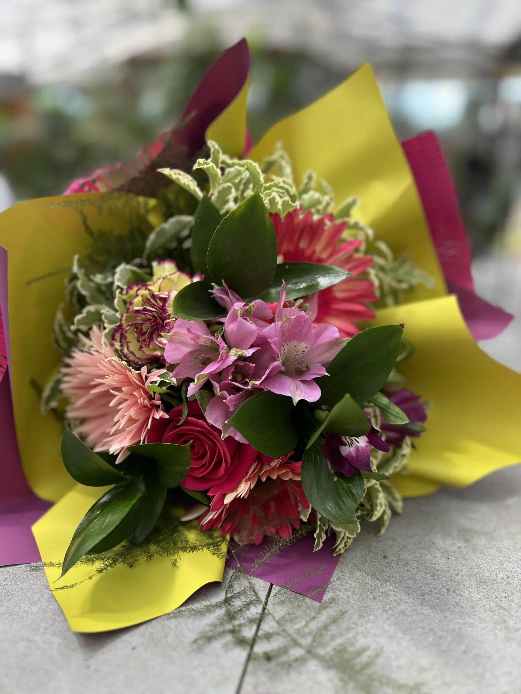 Mazzo di garofani, gerbera rosa e foglie variegate in carta gialla e viola, appoggiato sul banco