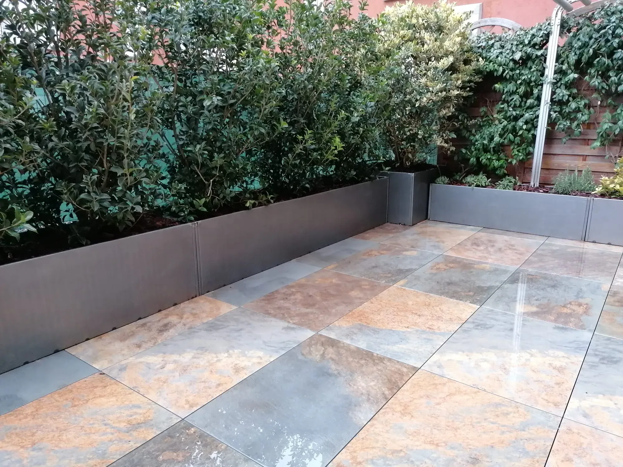 Terrazzo pavimentato con grandi lastre dalle sfumature ruggine e grigie, lucide di pioggia, con fioriere metalliche piene di arbusti lungo il bordo