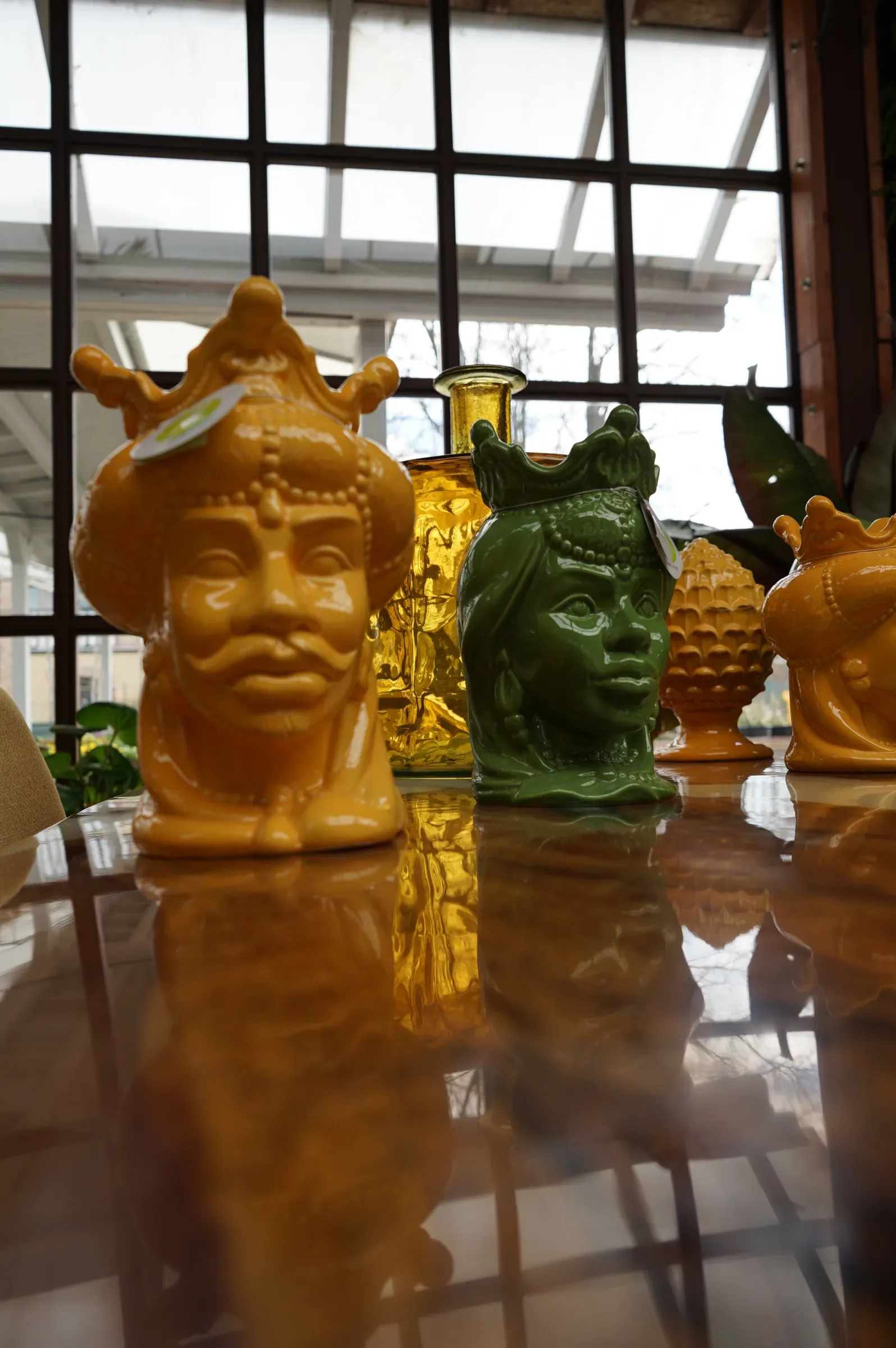 Vasi in ceramica gialla e verde a forma di testa con la corona, riflessi sul piano lucido di un tavolo