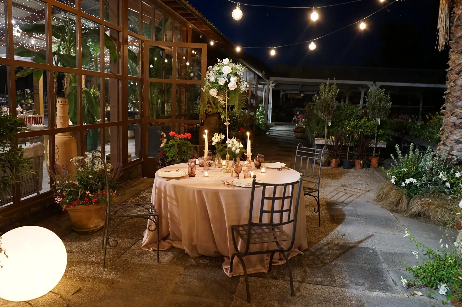 Tavola rotonda rosa con un'alzata di fiori bianchi fra le serre, sotto file di lampadine, con piante in vaso e una sfera luminosa