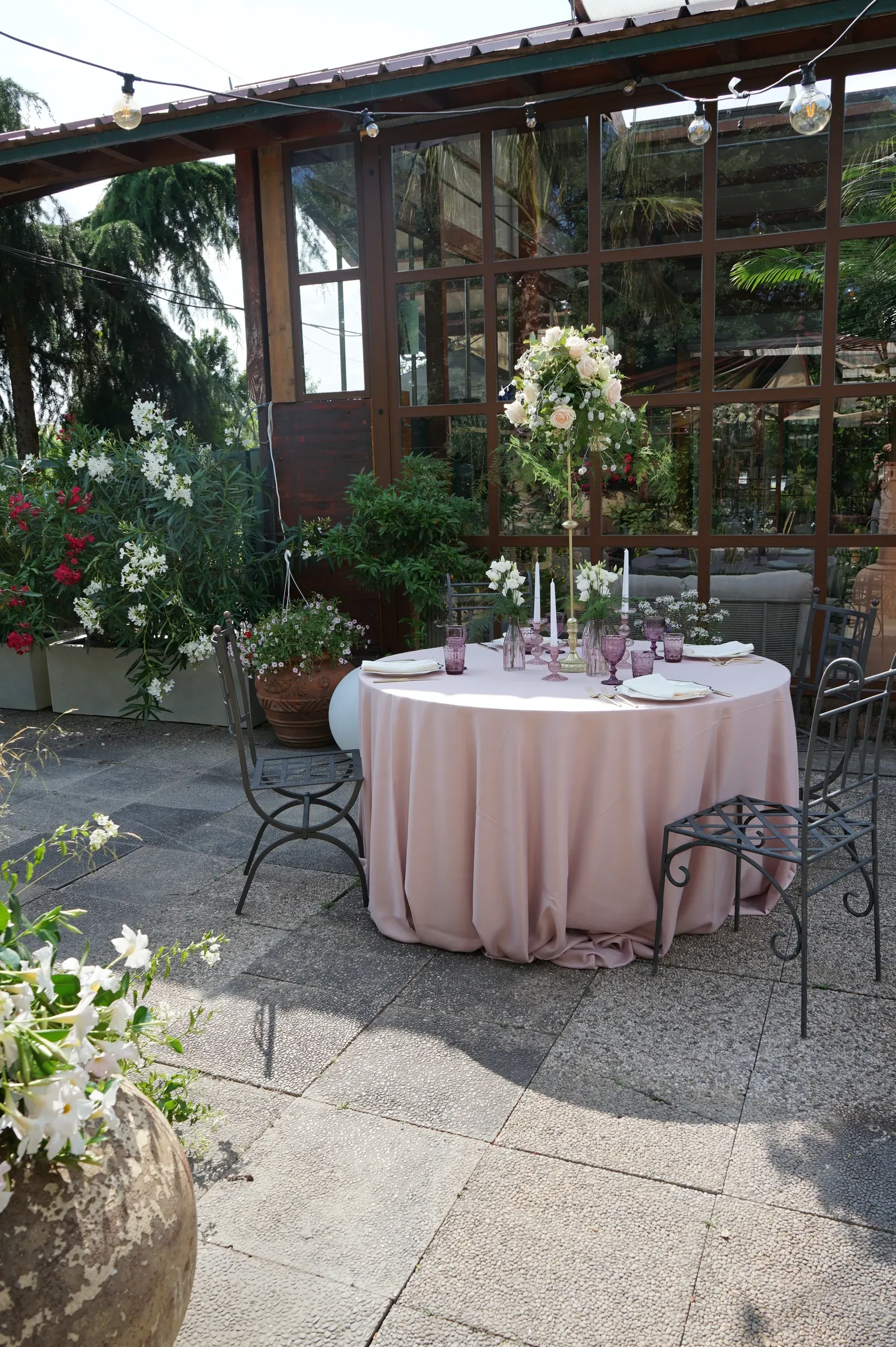Tavola rotonda rosa con un'alzata di fiori bianchi nel cortile davanti alla serra, con vasi di piante fiorite