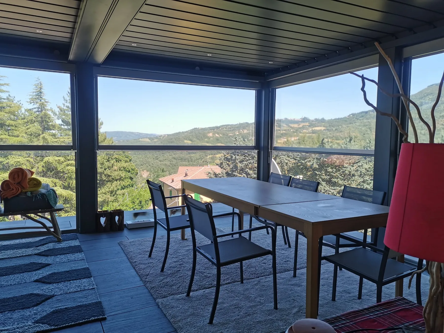 Pergola con le lamelle aperte e le pareti vetrate sulle colline, sopra un tavolo lungo su un pavimento scuro