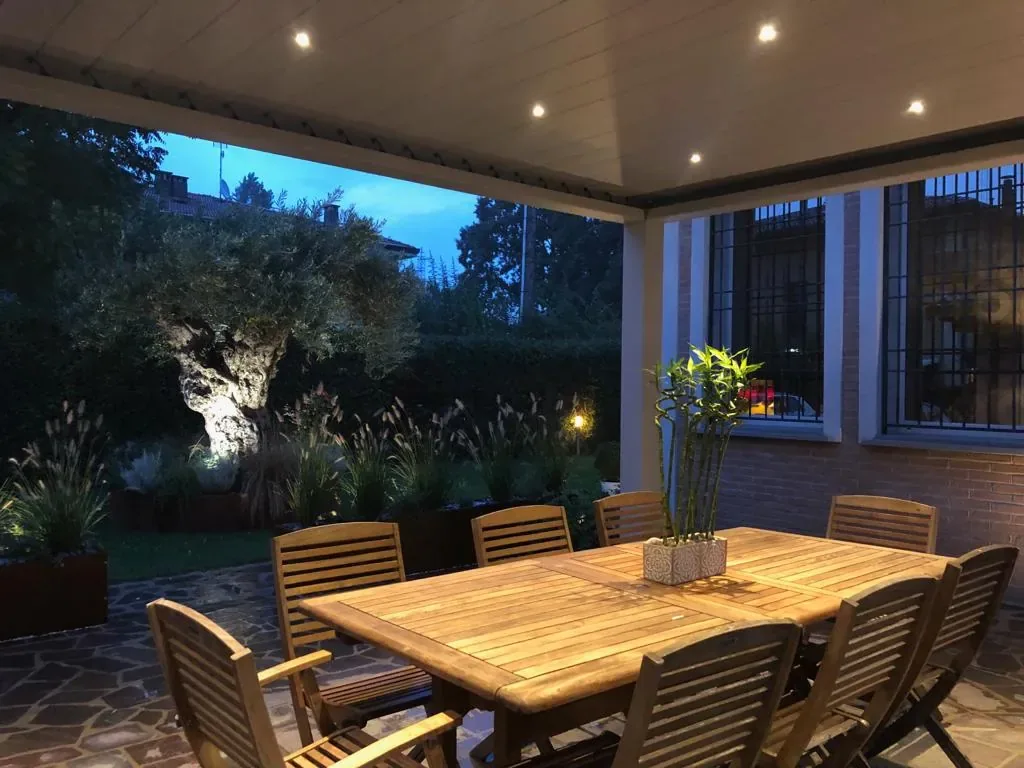 Pergola dal soffitto chiaro con i faretti accesi di sera, sopra un tavolo in teak con le sedie, e un olivo illuminato in giardino