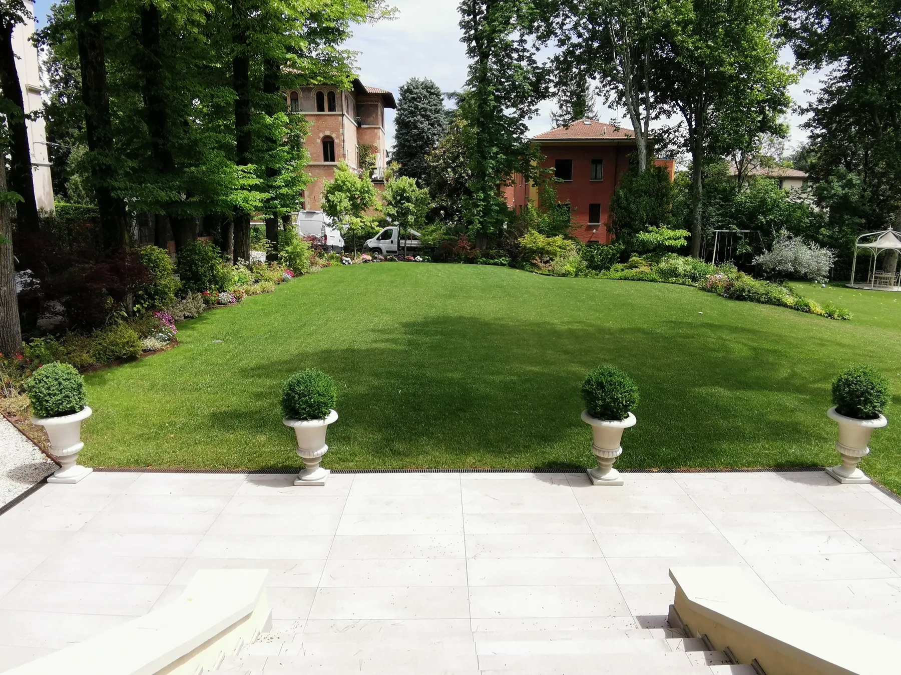 Grande prato rasato fra alberi alti, visto da una terrazza in pietra chiara con vasi di bosso allineati