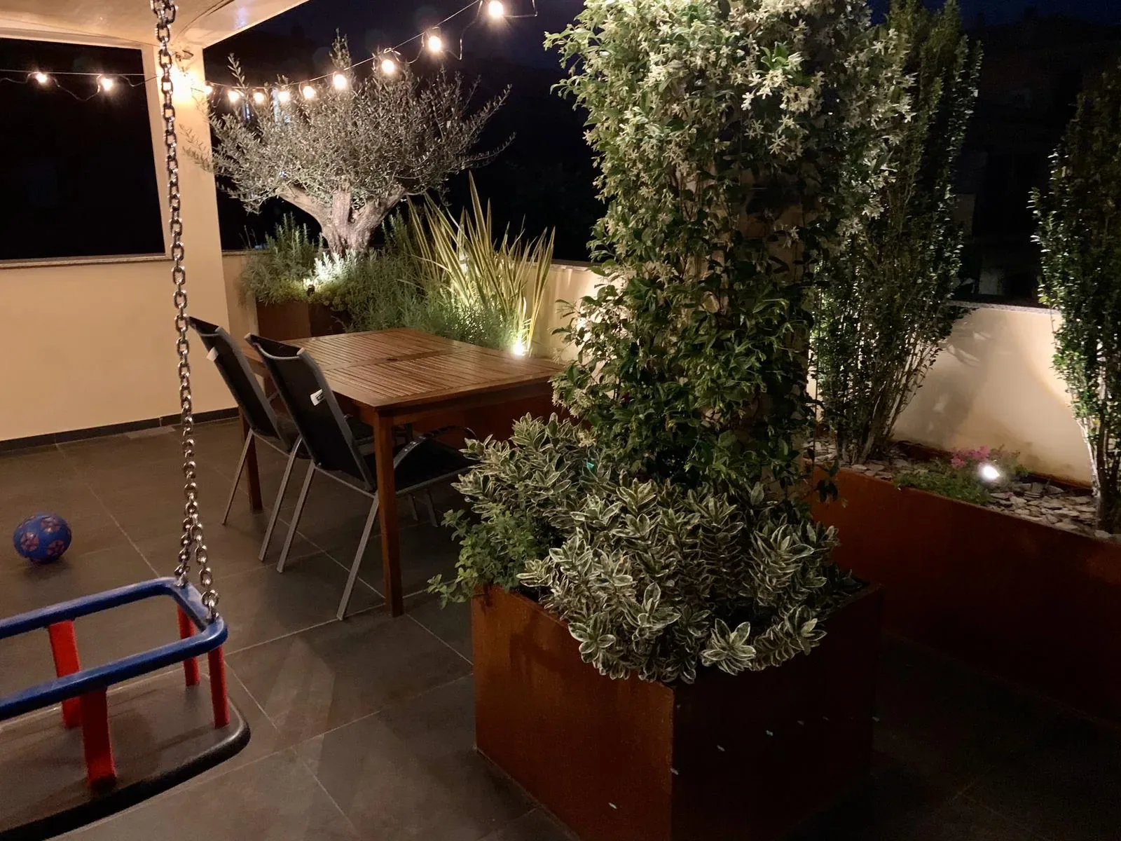 Terrazzo coperto di sera con un filo di lampadine, tavolo in legno e fioriere in corten con le piante illuminate dal basso