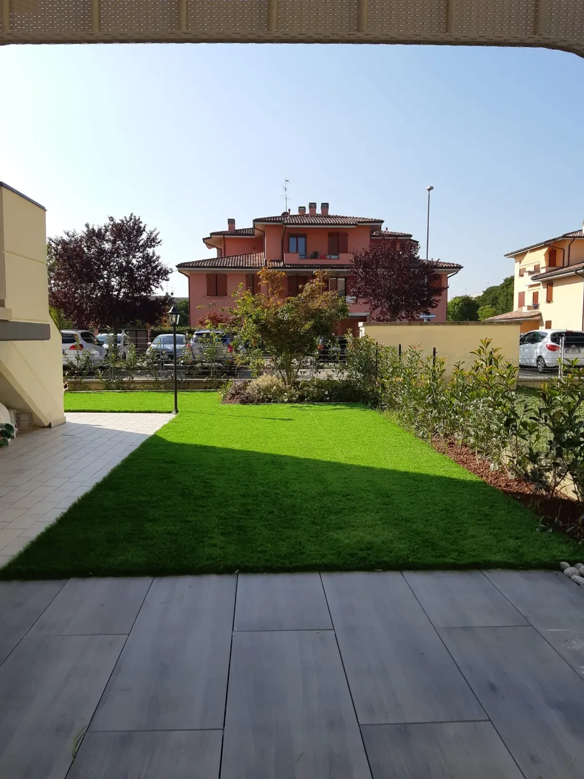 Prato sintetico stretto fra una pavimentazione effetto legno e un'aiuola di arbusti giovani, con alberi dalle foglie rosse e case sul fondo