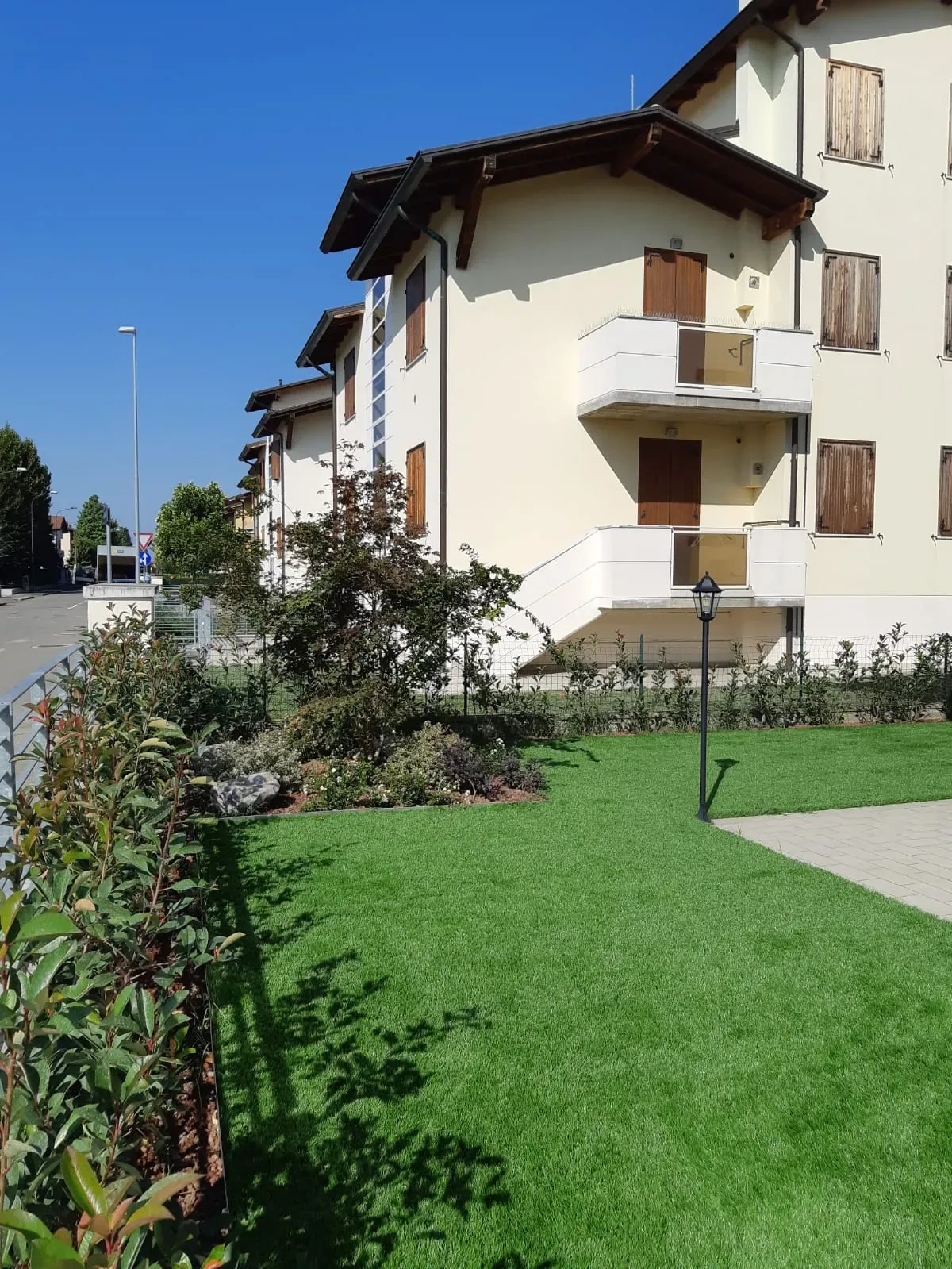 Prato sintetico ai piedi di una palazzina color crema con i balconi, bordato da un'aiuola di arbusti e da un lampione da giardino