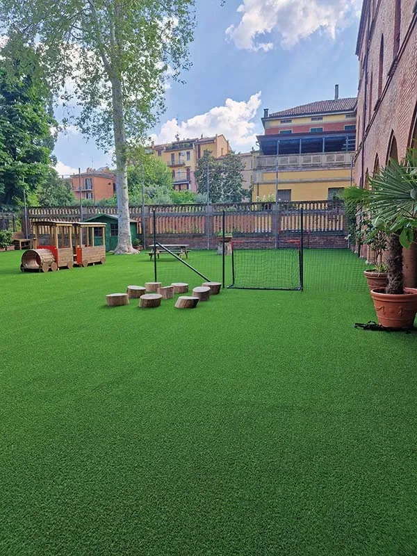 Cortile interno con prato sintetico, un trenino di legno, ceppi per sedersi e una porta da calcio, fra edifici in mattoni