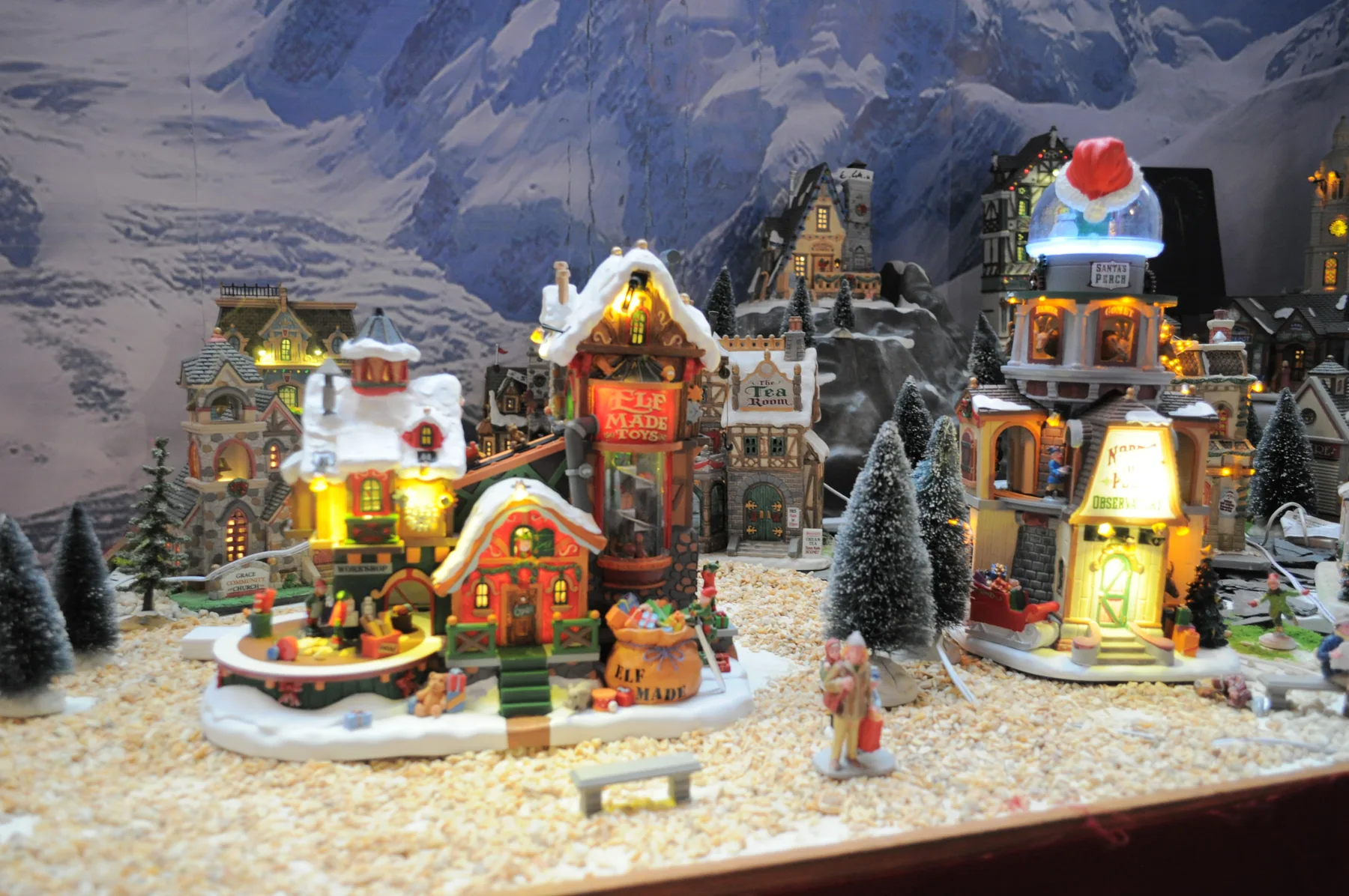 Villaggio in miniatura con case illuminate e alberelli innevati, davanti a un fondale di montagne