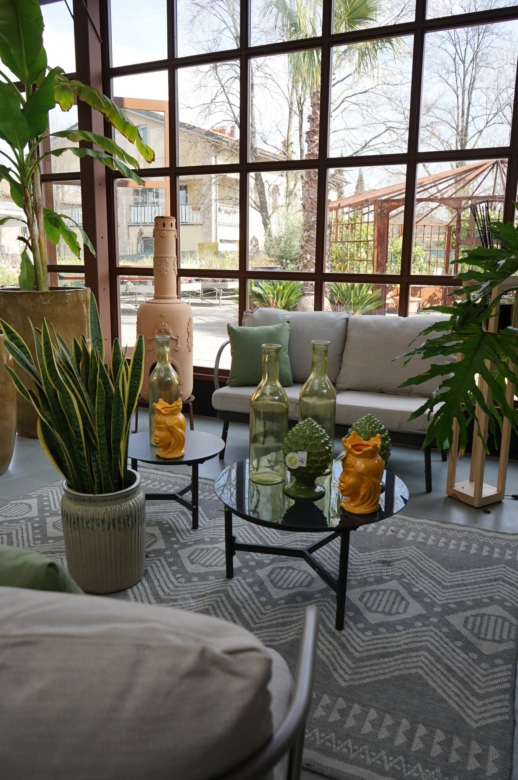 Salotto sotto la serra vetrata con divano chiaro, sansevieria in vaso e bottiglie di vetro sul tavolino