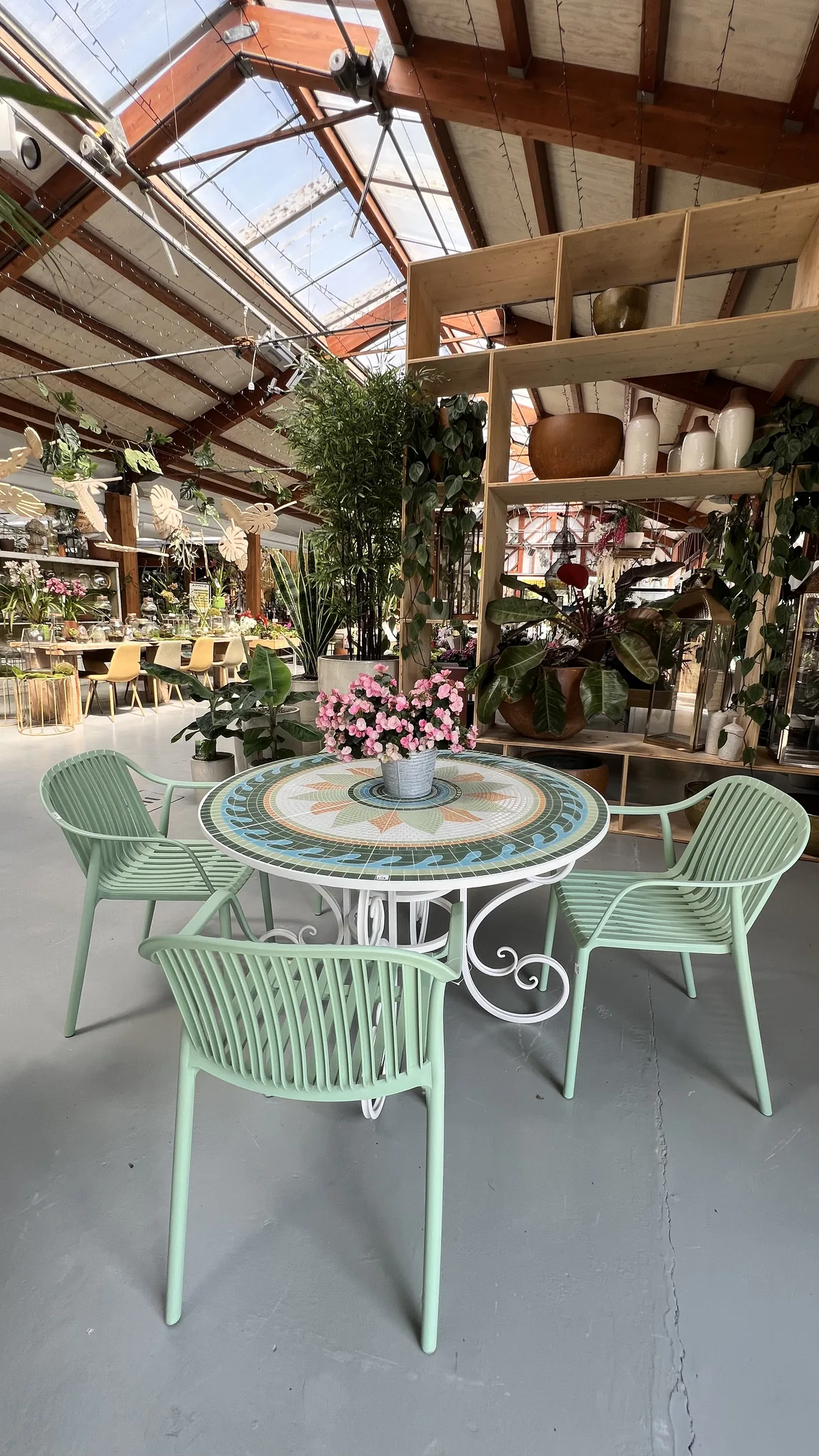 Tavolo tondo con il piano in mosaico e sedie verde menta sotto la serra, con una composizione di fiori rosa al centro e gli scaffali di vasi sul fondo