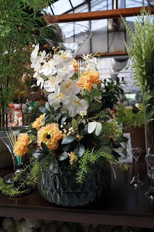 Composizione artificiale di orchidee bianche, garofani arancioni e felci in un vaso di vetro scuro