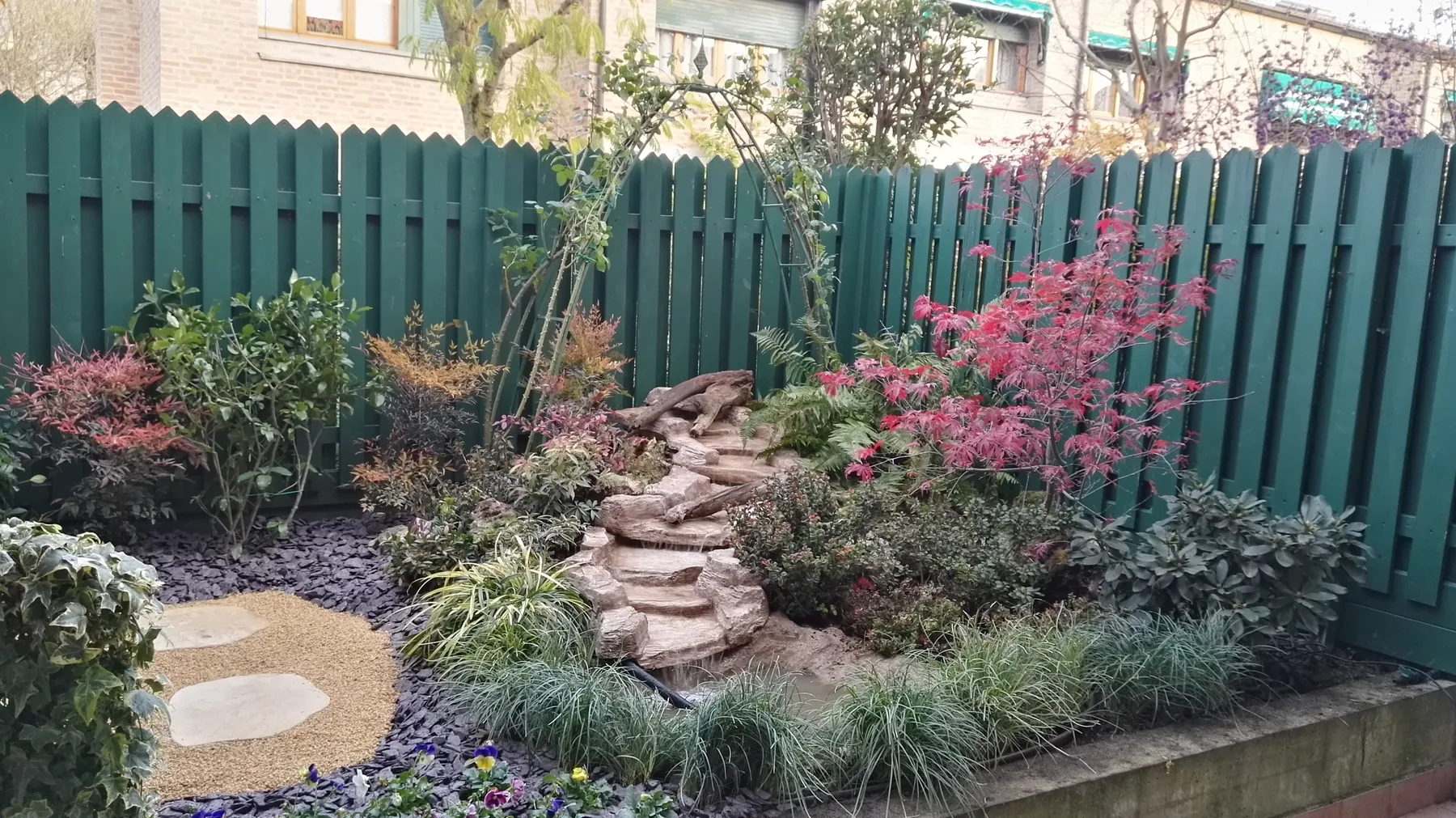 Aiuola contro uno steccato verde, con aceri, arbusti dal fogliame colorato e una piccola cascata di pietre