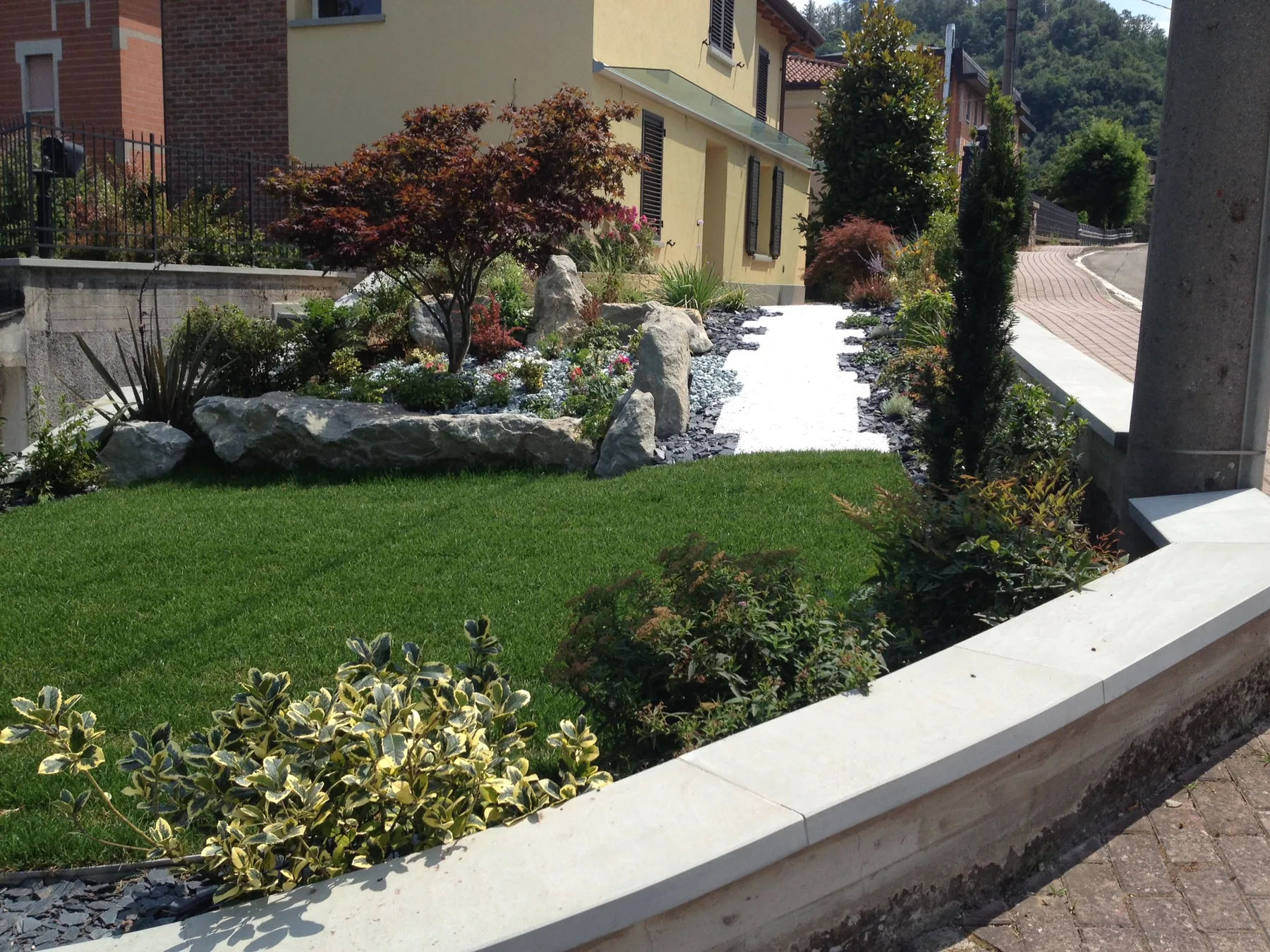 Prato in pendenza chiuso da un muretto, con aceri rossi, massi e arbusti lungo il vialetto di una casa
