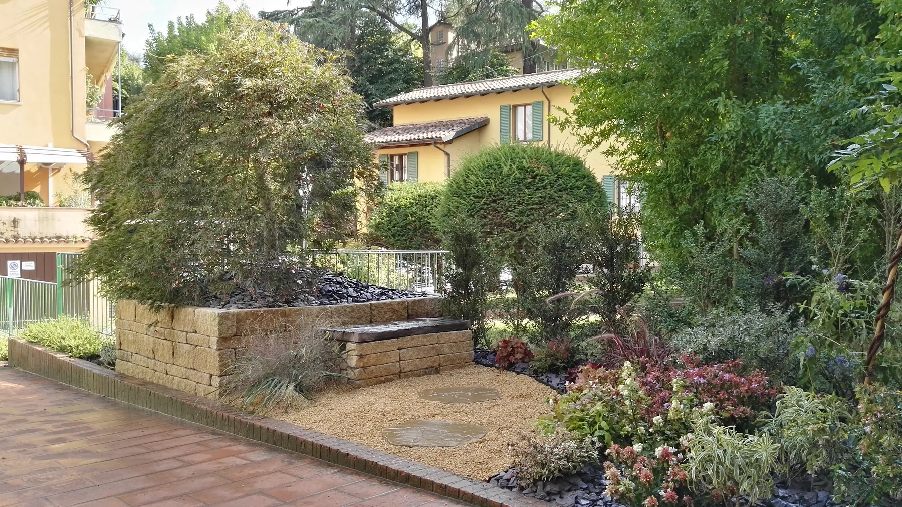 Aiuola rialzata in pietra con un acero, ghiaia, arbusti potati e cespugli fioriti lungo il marciapiede di un condominio